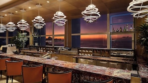 A modern kitchen with a marble countertop, pendant lights, and a view of the sunset through large windows.