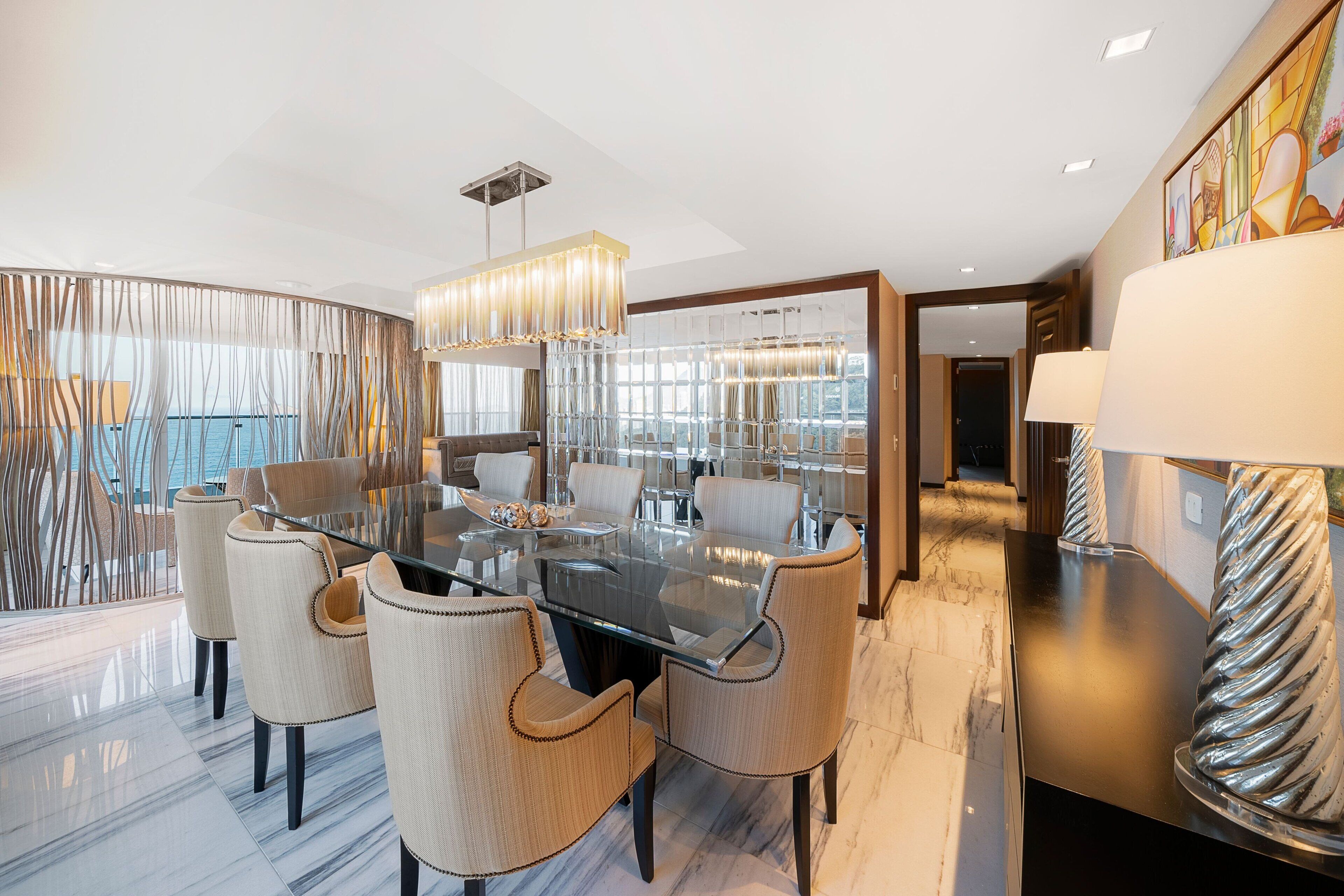 Uma sala de jantar moderna com mesa de vidro rodeada por cadeiras estofadas, um lustre contemporâneo e vista da cidade através de grandes janelas.