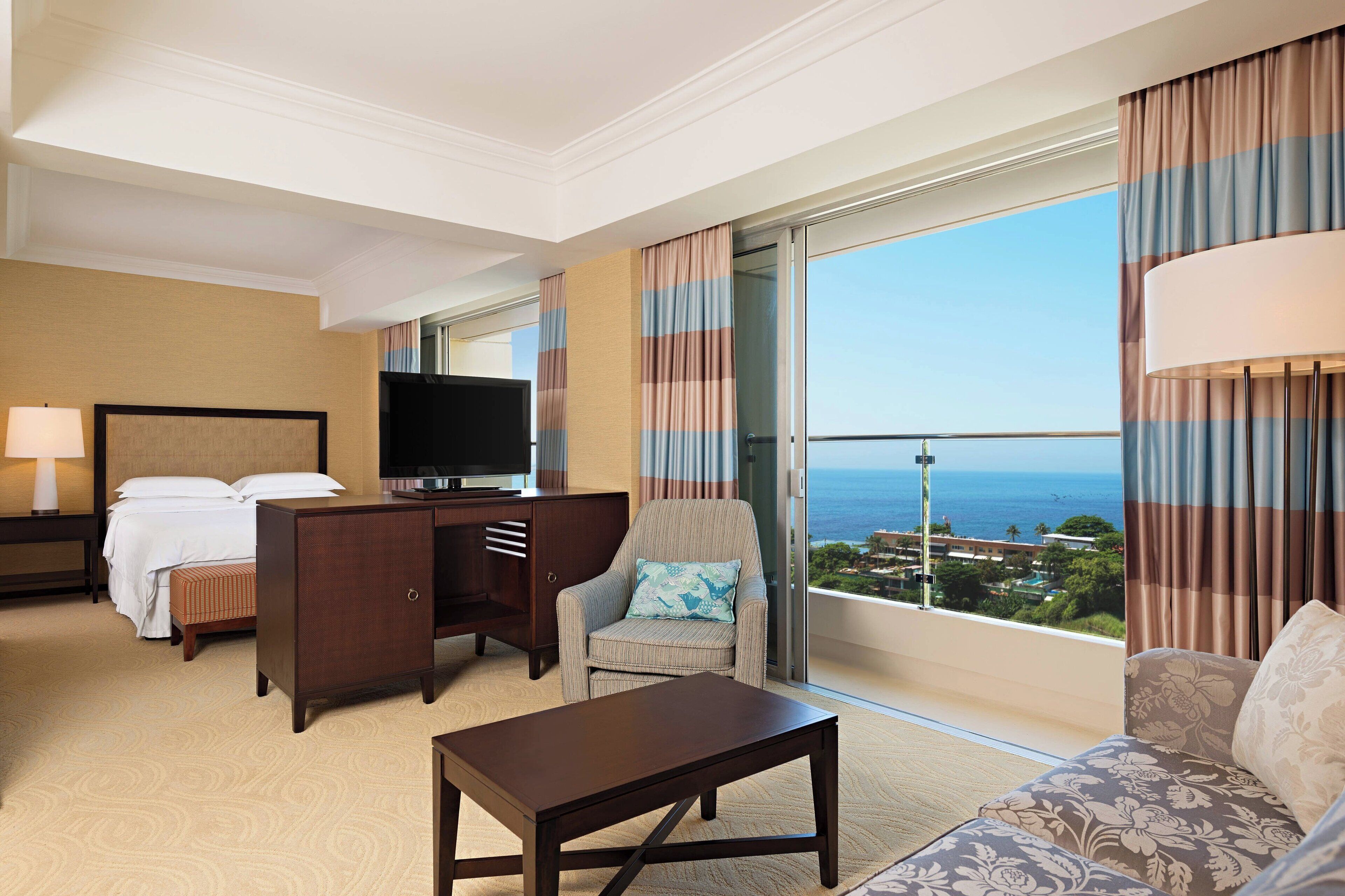 A hotel room with a bed, a TV, a desk, a chair, a sofa, and a large window with a view of the ocean.