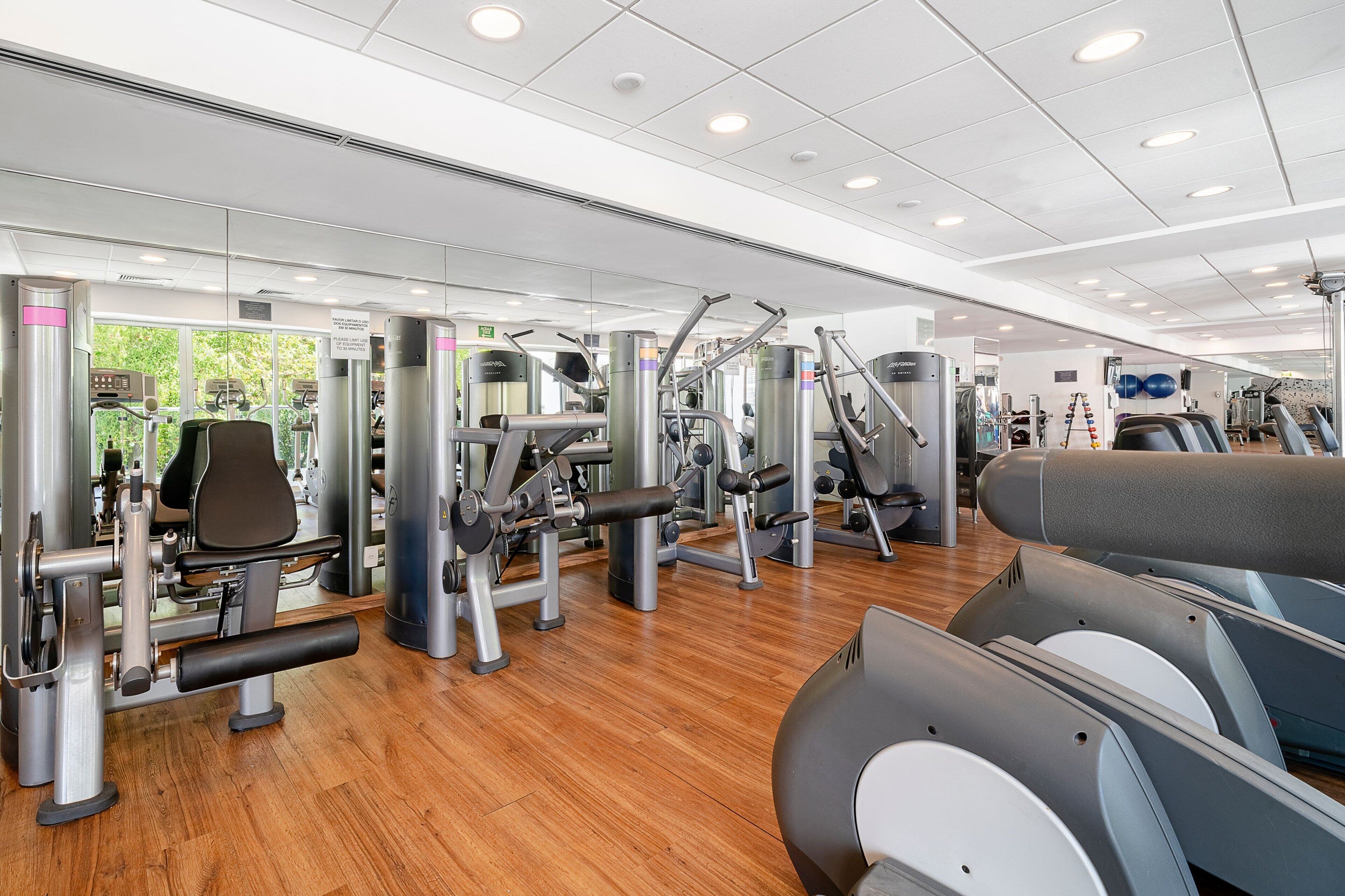 Academia moderna com diversos equipamentos de exercício, piso de madeira e amplas janelas.