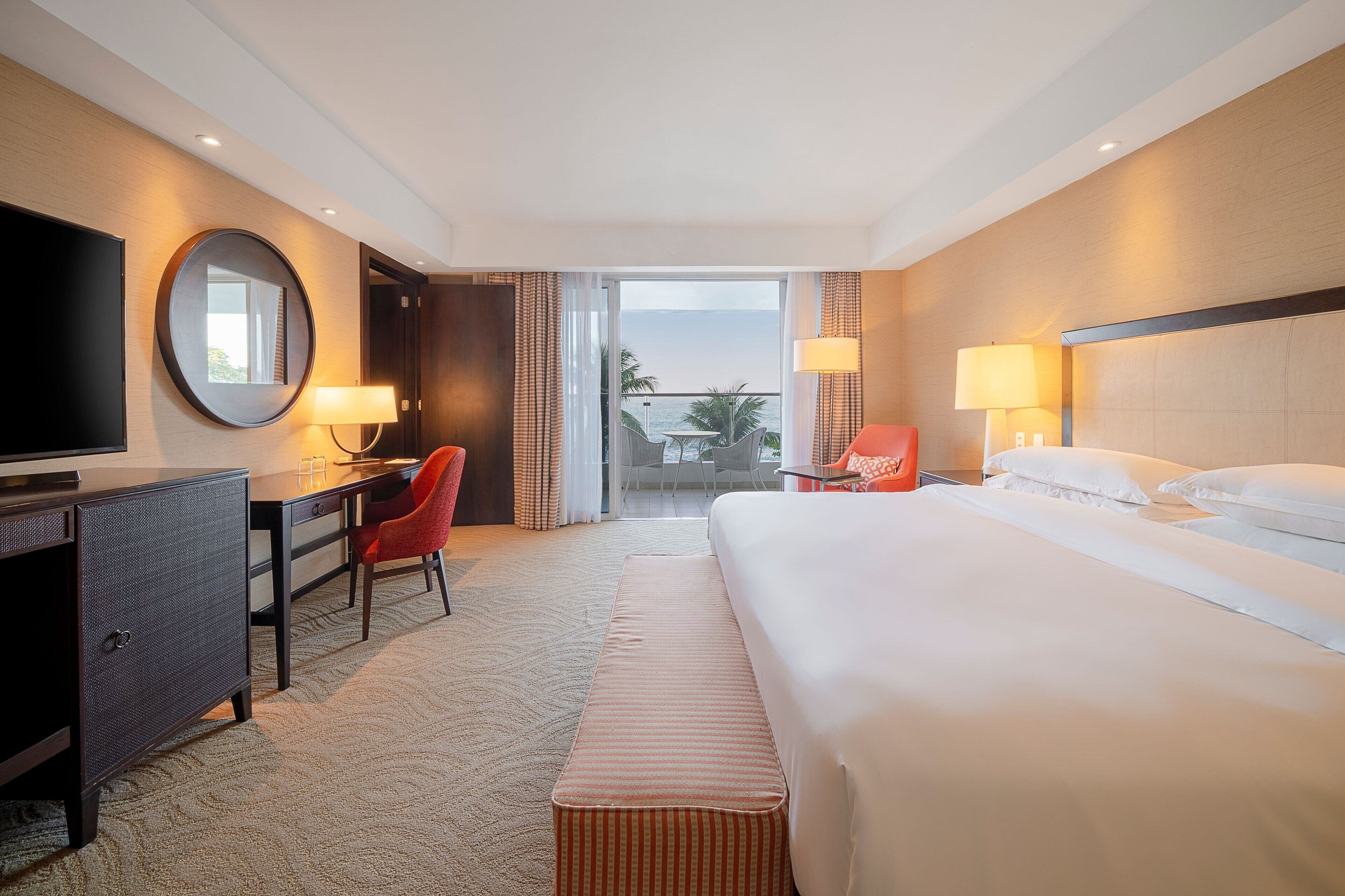 Ein Hotelzimmer mit Bett, Schreibtisch mit Stuhl, Fernseher und einem Balkon mit Meerblick.