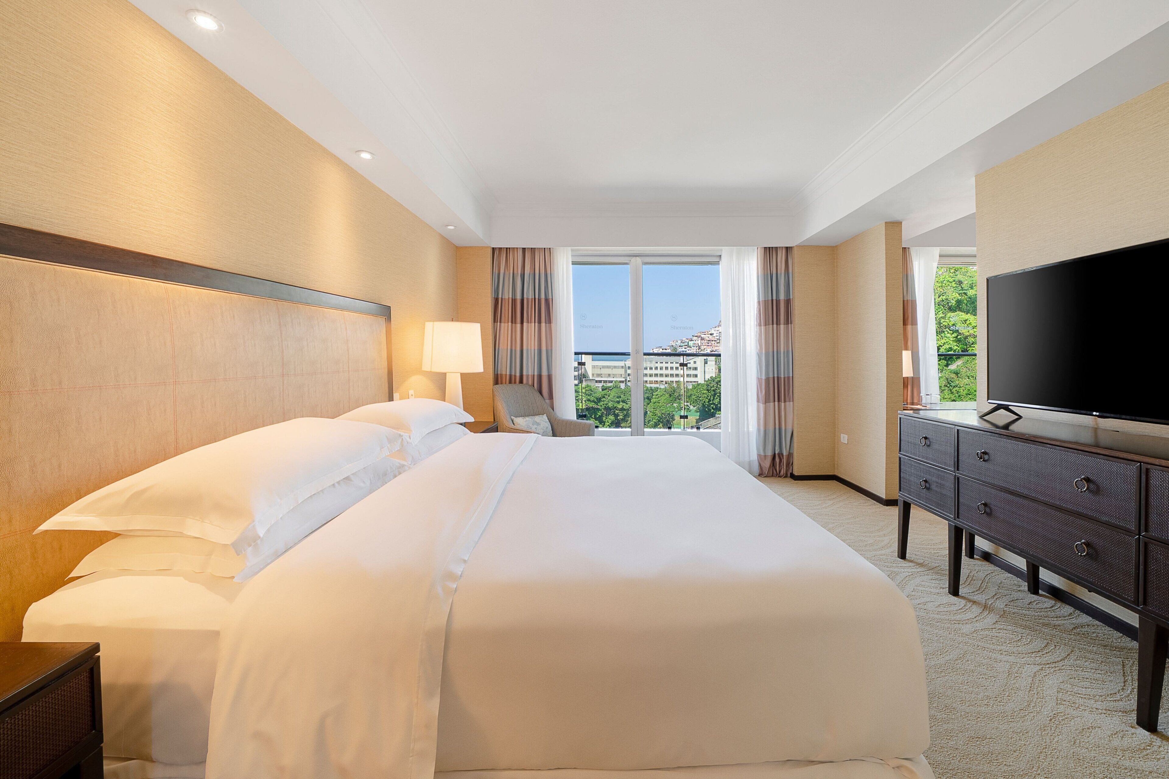 Habitación de hotel con una cama grande, televisión, cómoda y vistas al exterior.
