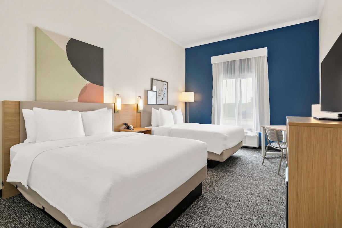 A hotel room with two beds, a desk, a chair, and a large window with curtains.