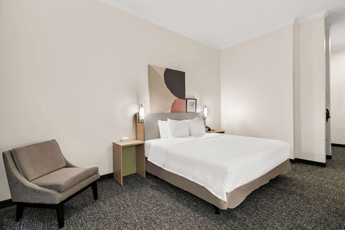 A hotel room with a bed, a chair, a nightstand, and a wall-mounted artwork.