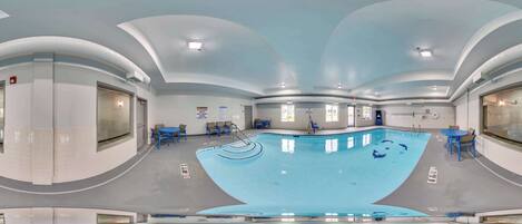 Una amplia piscina interior con techo curvo, rodeada de sillas y mesas azules, y vistas claras al área de la piscina.