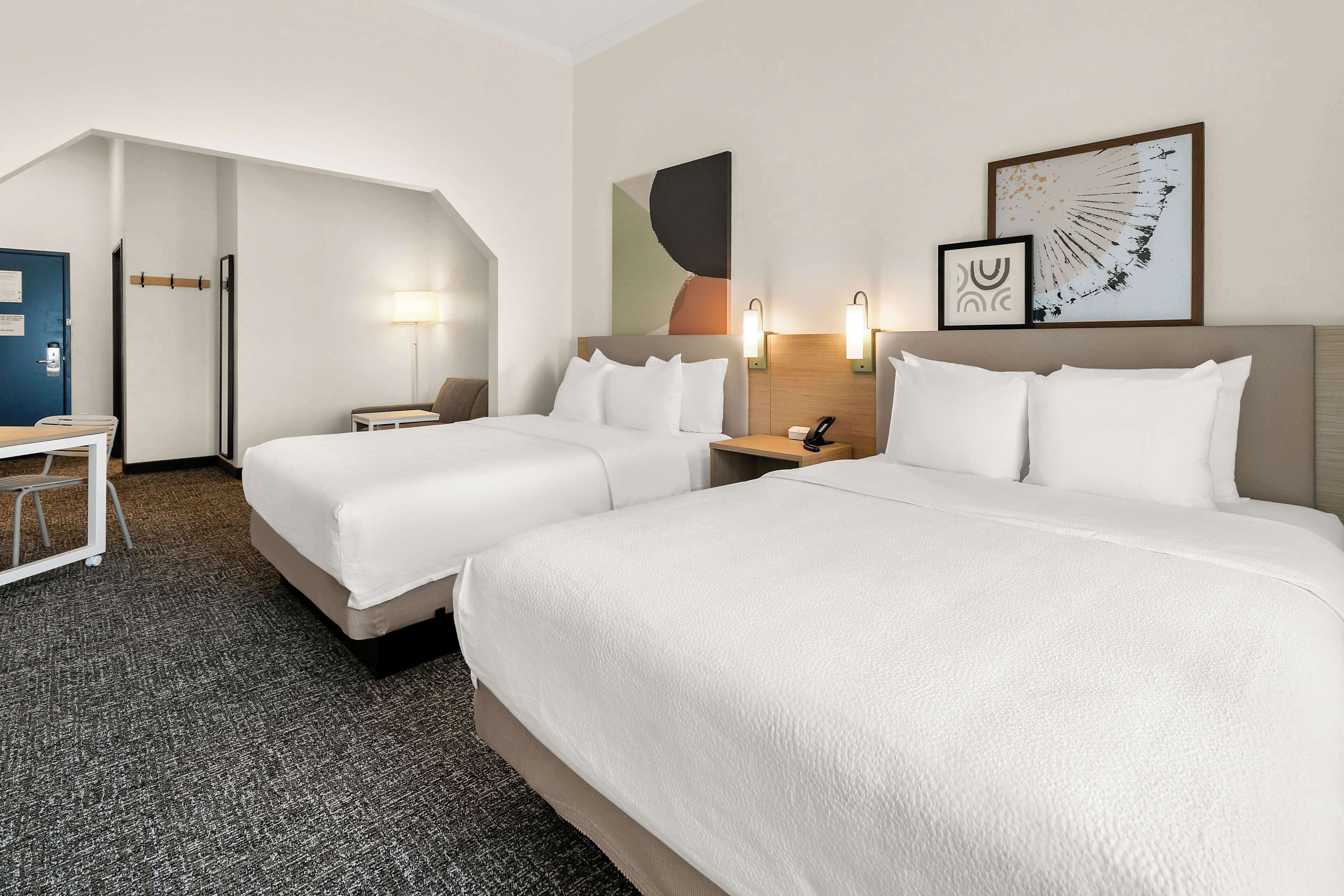 Habitación de hotel con dos camas, un escritorio y una silla. Hay lámparas de pared y una obra de arte enmarcada sobre las camas.