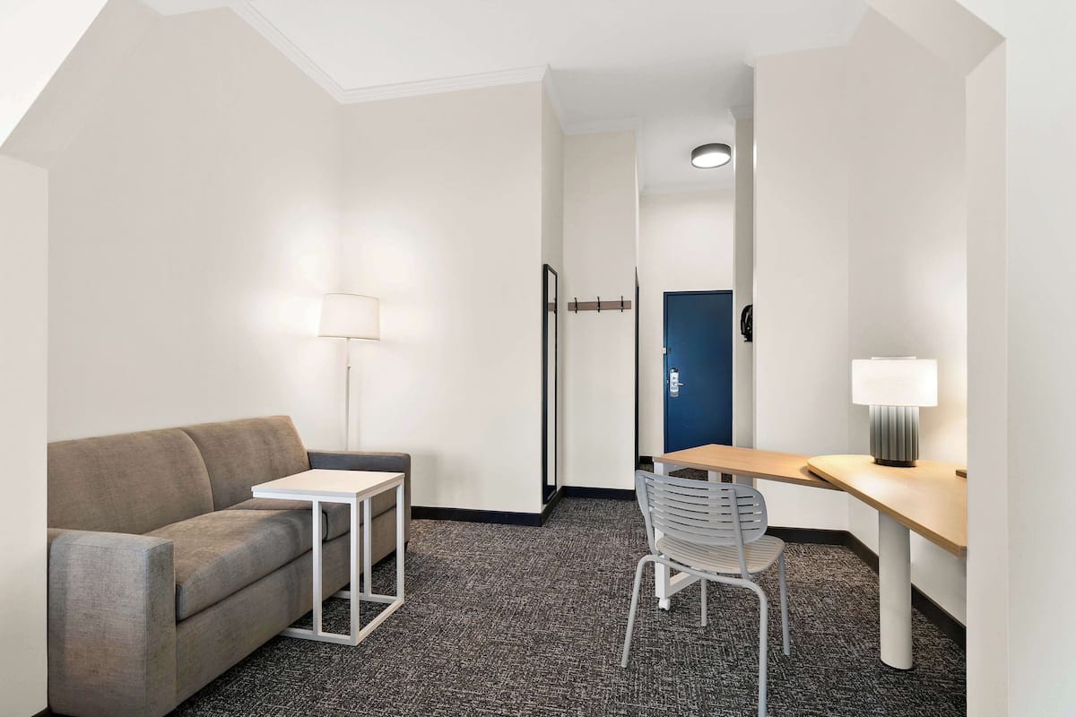 A hotel room with a gray sofa, a small white table, a chair, a desk, and a lamp.
