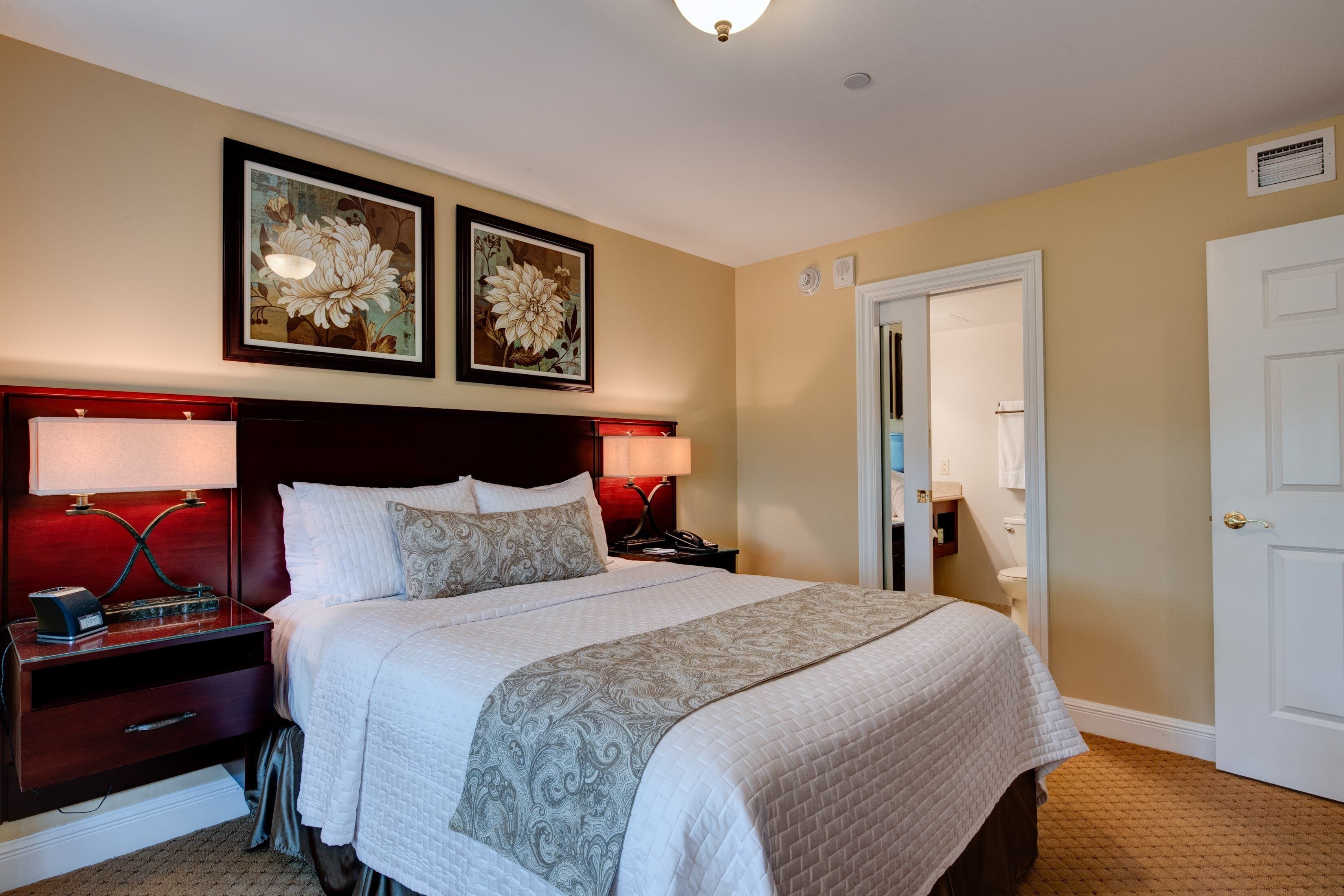 A neatly made bed with a white and gray patterned bedspread, two framed floral paintings above it, a red bedside lamp, and a white door to the right.