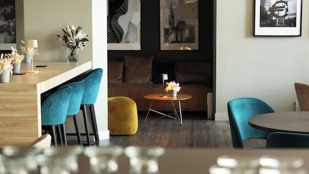 Un espace repas moderne avec un bar, des chaises bleues, un pouf jaune et des œuvres d’art encadrées accrochées aux murs.