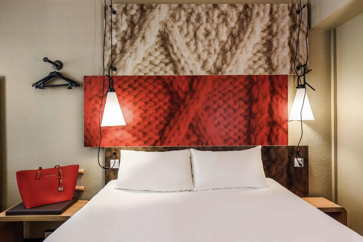 Een hotelkamer met een bed, witte kussens, een rode handtas en een hoofdbord met rode en beige textuur.
