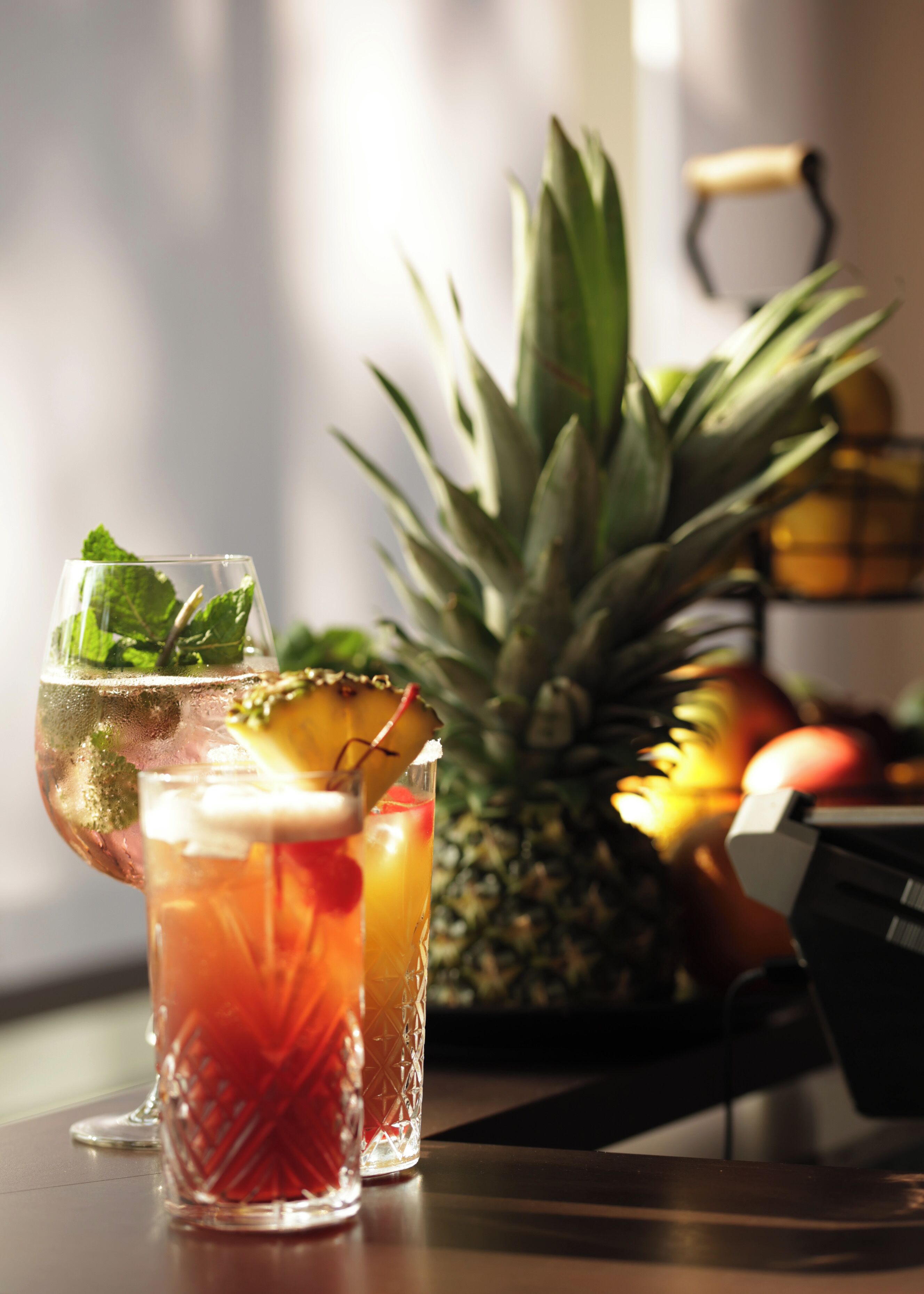 Two glasses of a red beverage with garnishes, a pineapple, and a bottle of orange juice on a table.