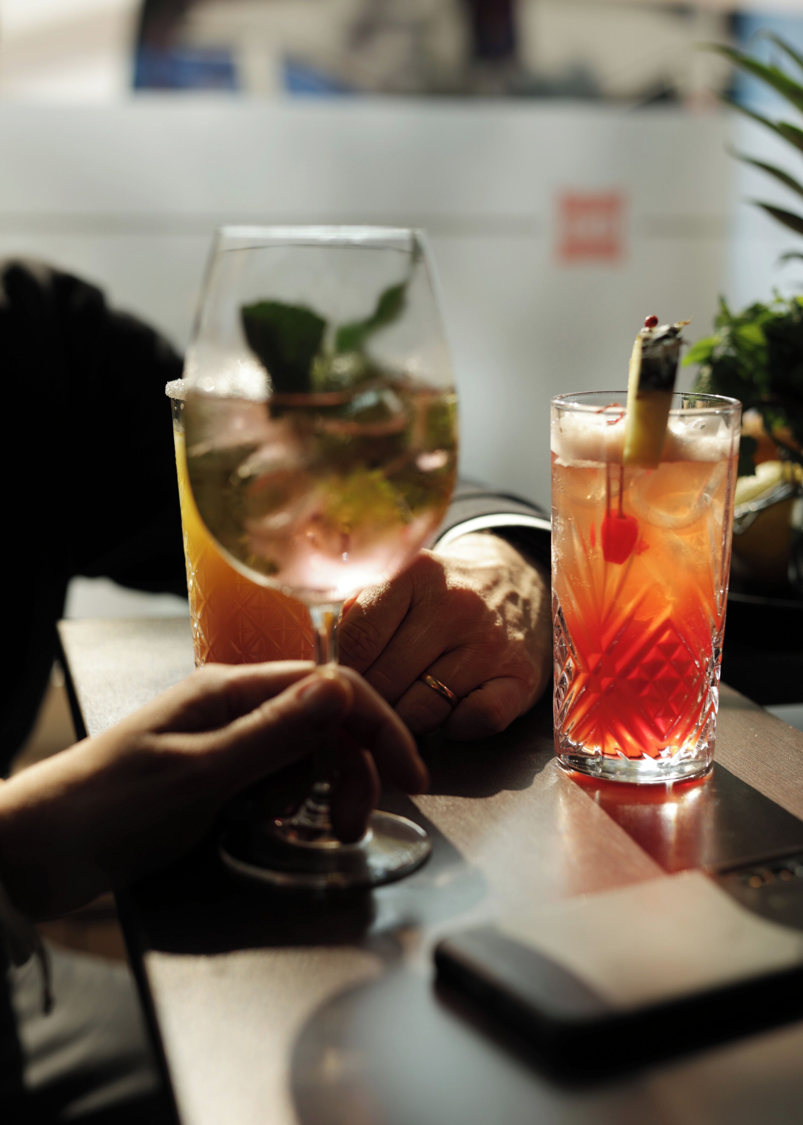 Two glasses of beverages on a table with a smartphone and a plant in the background.