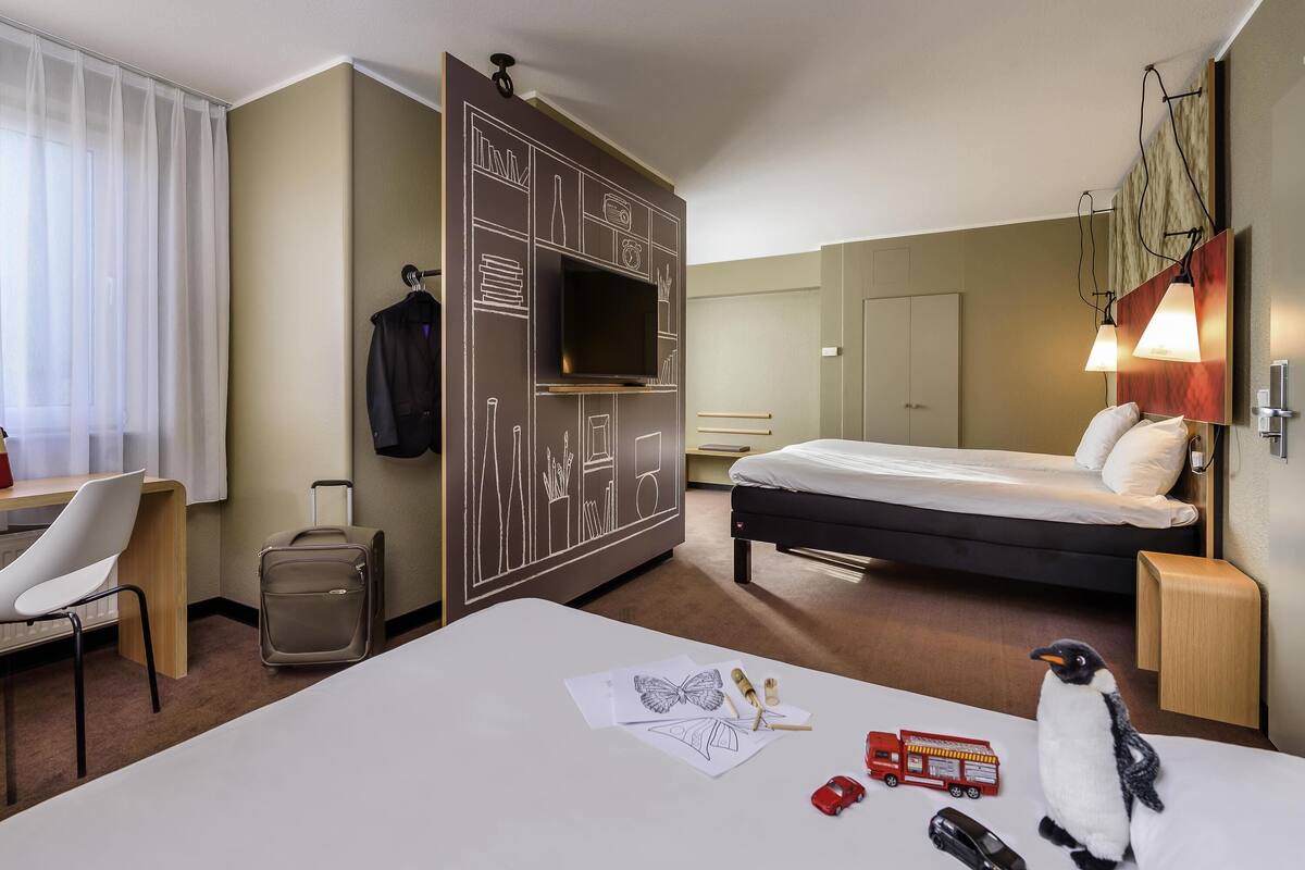 Een hotelkamer met een bed, bureau, stoel, kledingkast en een kleine tafel met een pluchen pinguïn.