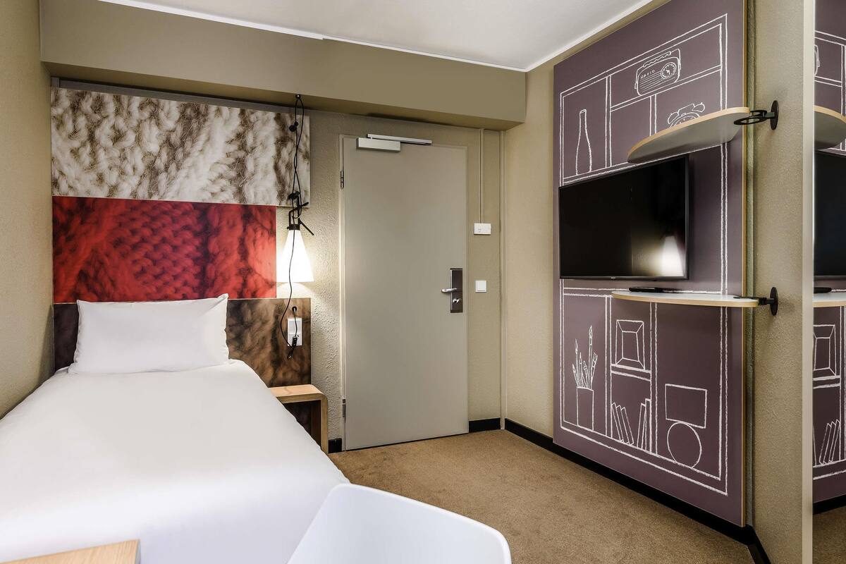 Een hotelkamer met een bed, nachtkastje, lamp, televisie en een garderobe met krijtborddecoraties.