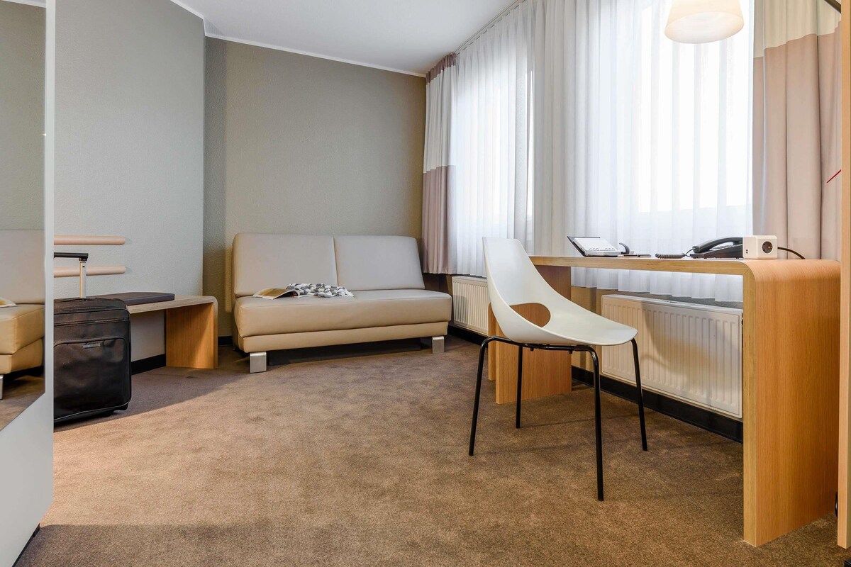 Una habitación de hotel moderna con sofá, escritorio con computadora portátil, silla, maleta y radiador.