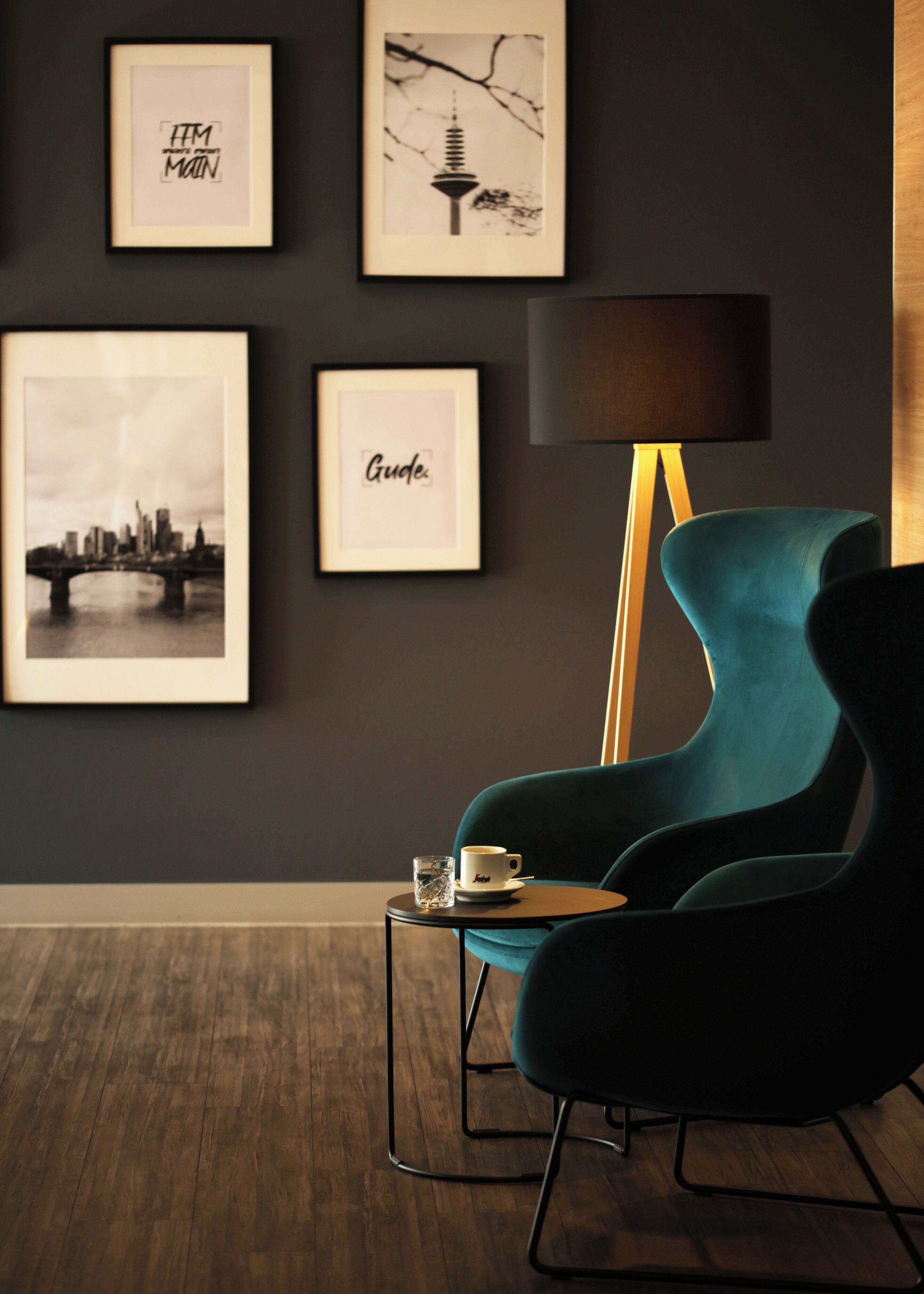 A modern room with a teal armchair, a coffee table with a cup and glass, and framed pictures on the wall.