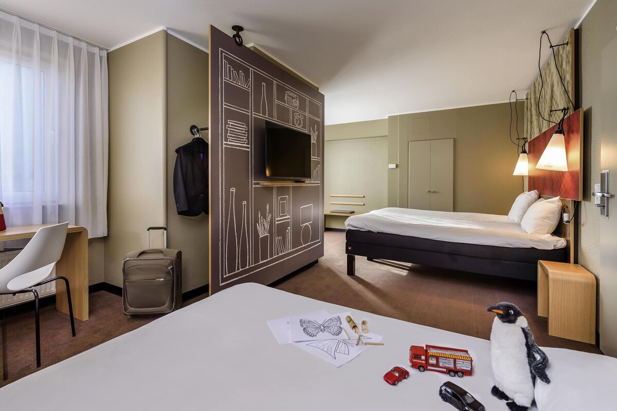 Een hotelkamer met een bed, bureau, stoel, kledingkast en een kleine tafel met een pluchen pinguïn.