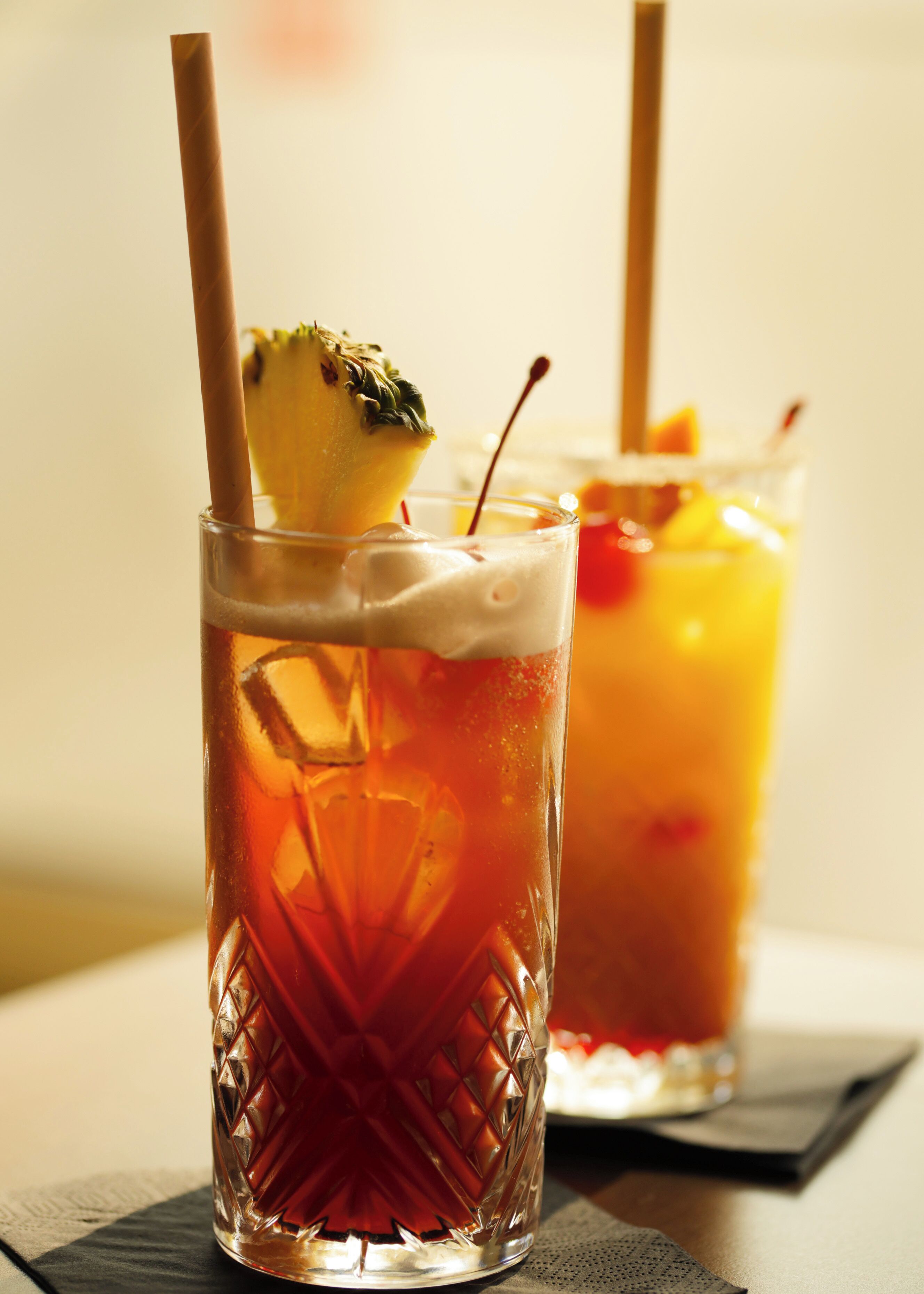 Twee glazen met ijskoude drankjes, rietjes en garnering, geplaatst op een tafel met een servet.