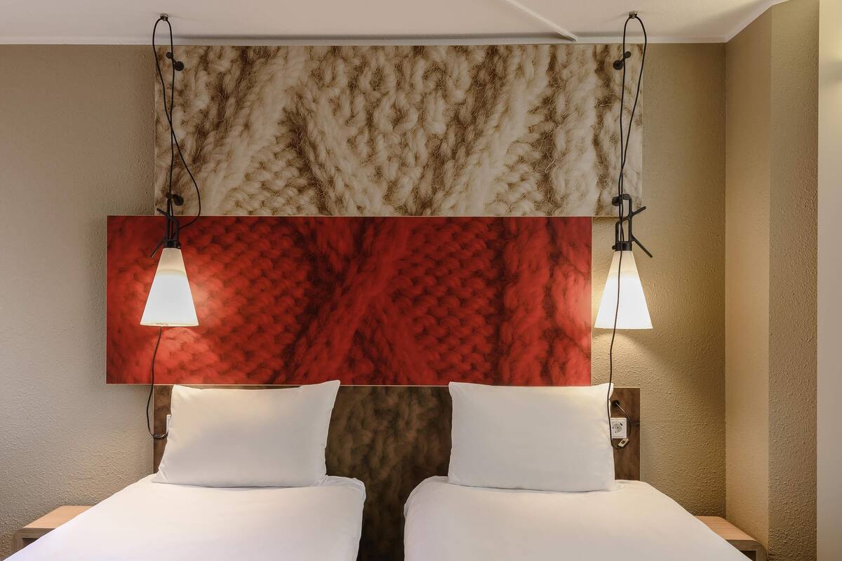 Een hotelkamer met twee bedden, een wand met rode en bruine textuur, en twee hanglampen.