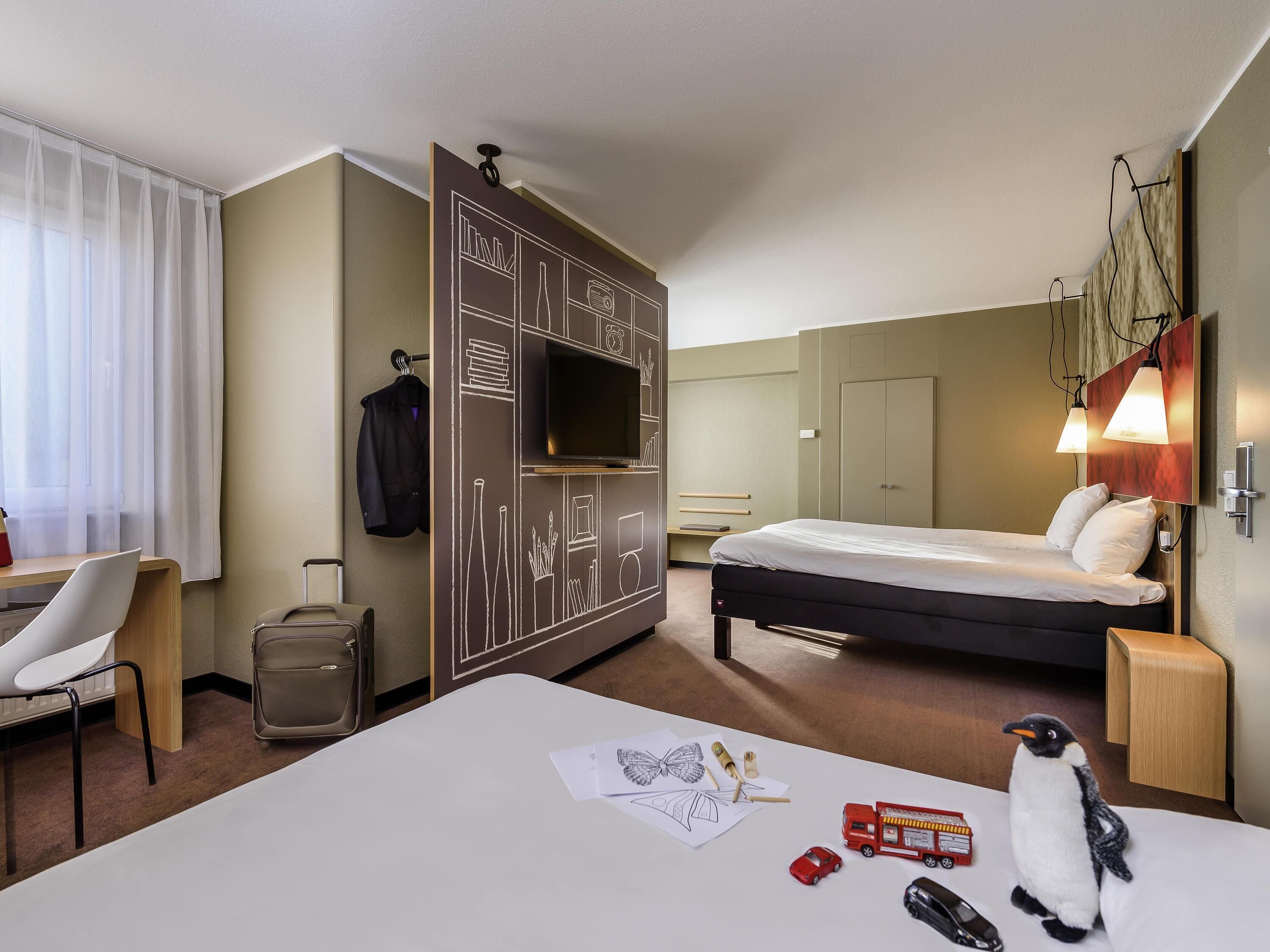 A hotel room with a bed, desk, chair, wardrobe, and a small table with a penguin plush toy.