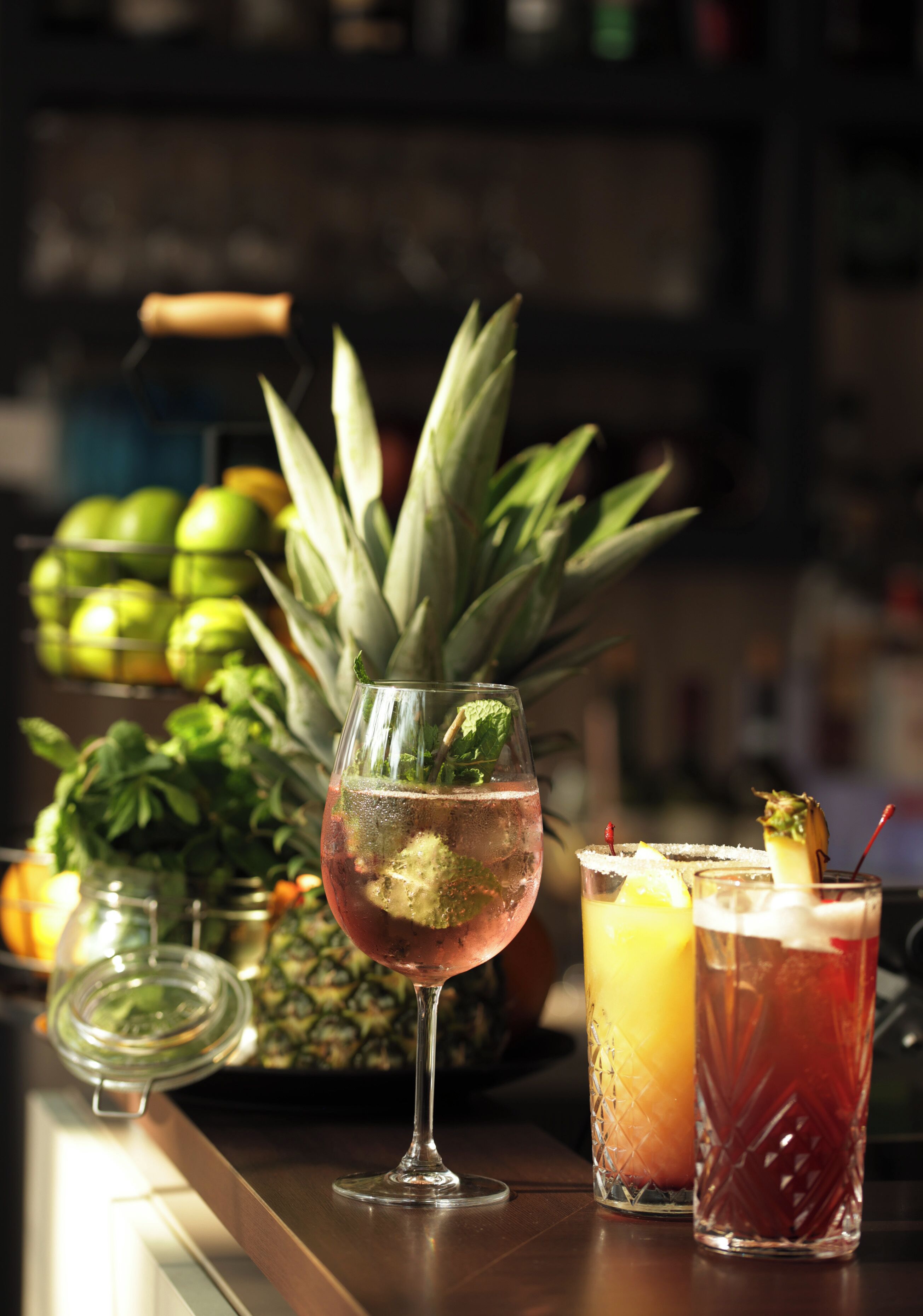 A bar setting with a glass of pink cocktail, a pineapple, and two other drinks on the counter.