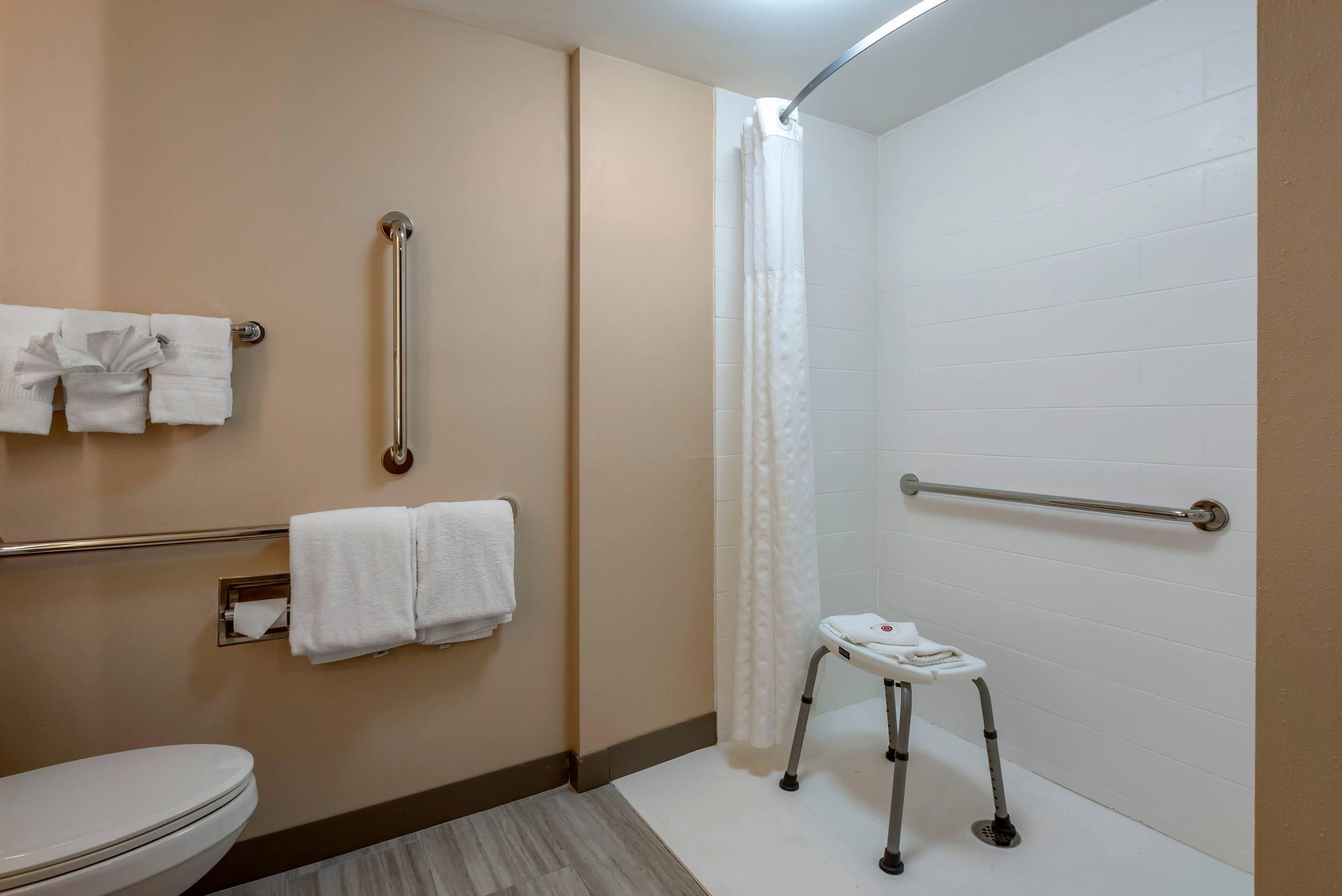 Een badkamer met toilet, twee handdoekrekken, een greep, een douchegordijn en een rolstoeltoegankelijke bank.