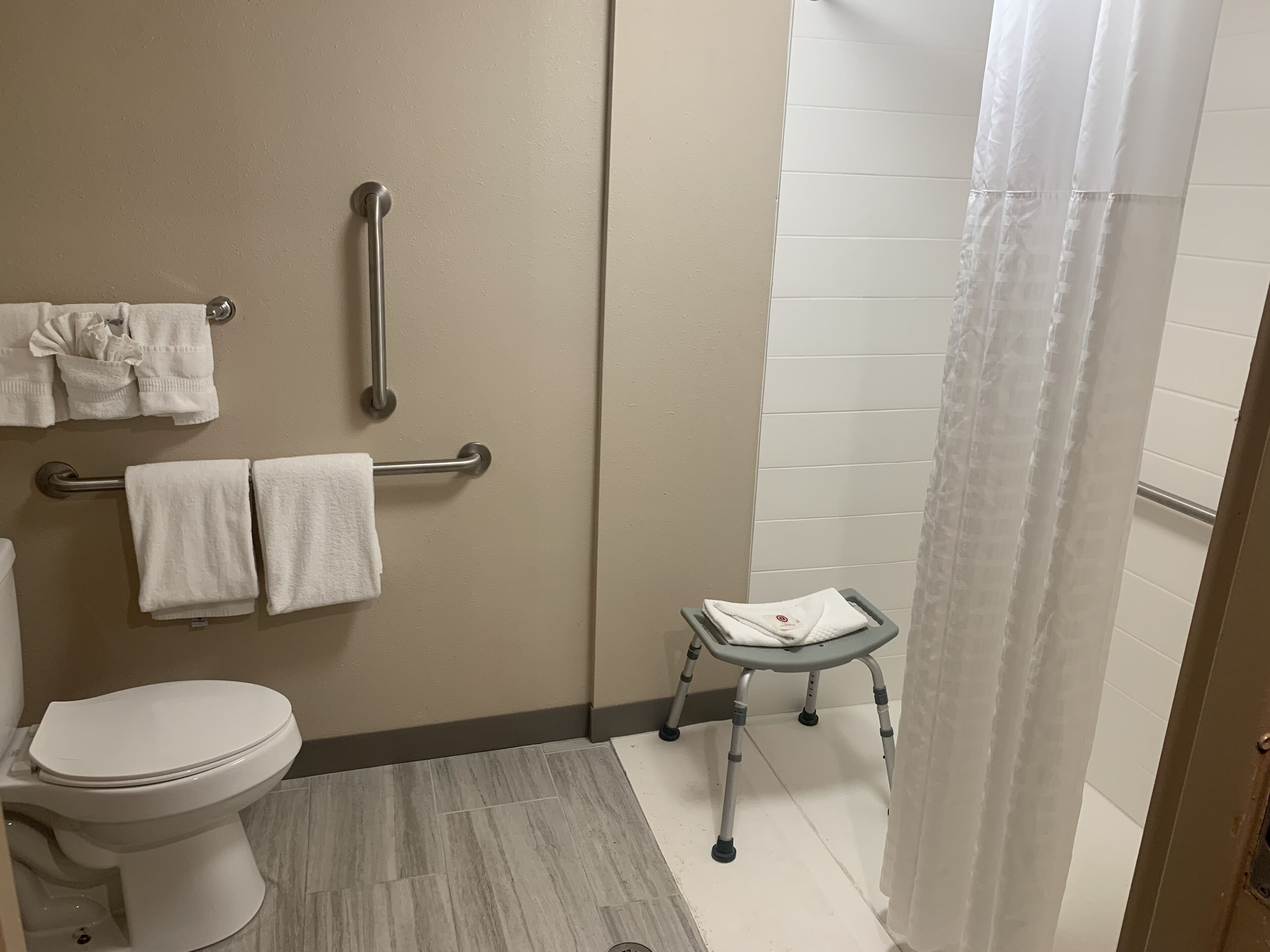 Baño con inodoro, barras de apoyo, cortina de ducha y un taburete pequeño.