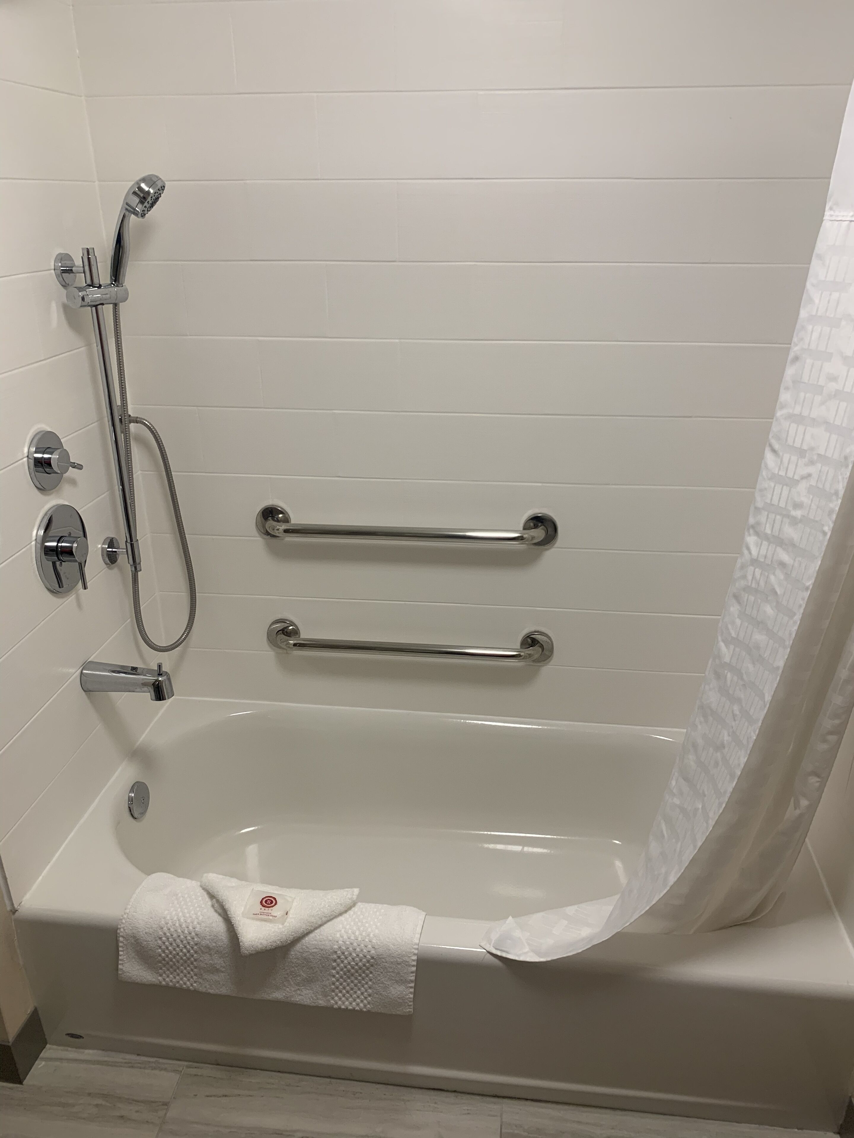 Un baño moderno con una bañera blanca, barras de apoyo, un cabezal de ducha manual y un perchero para toallas.