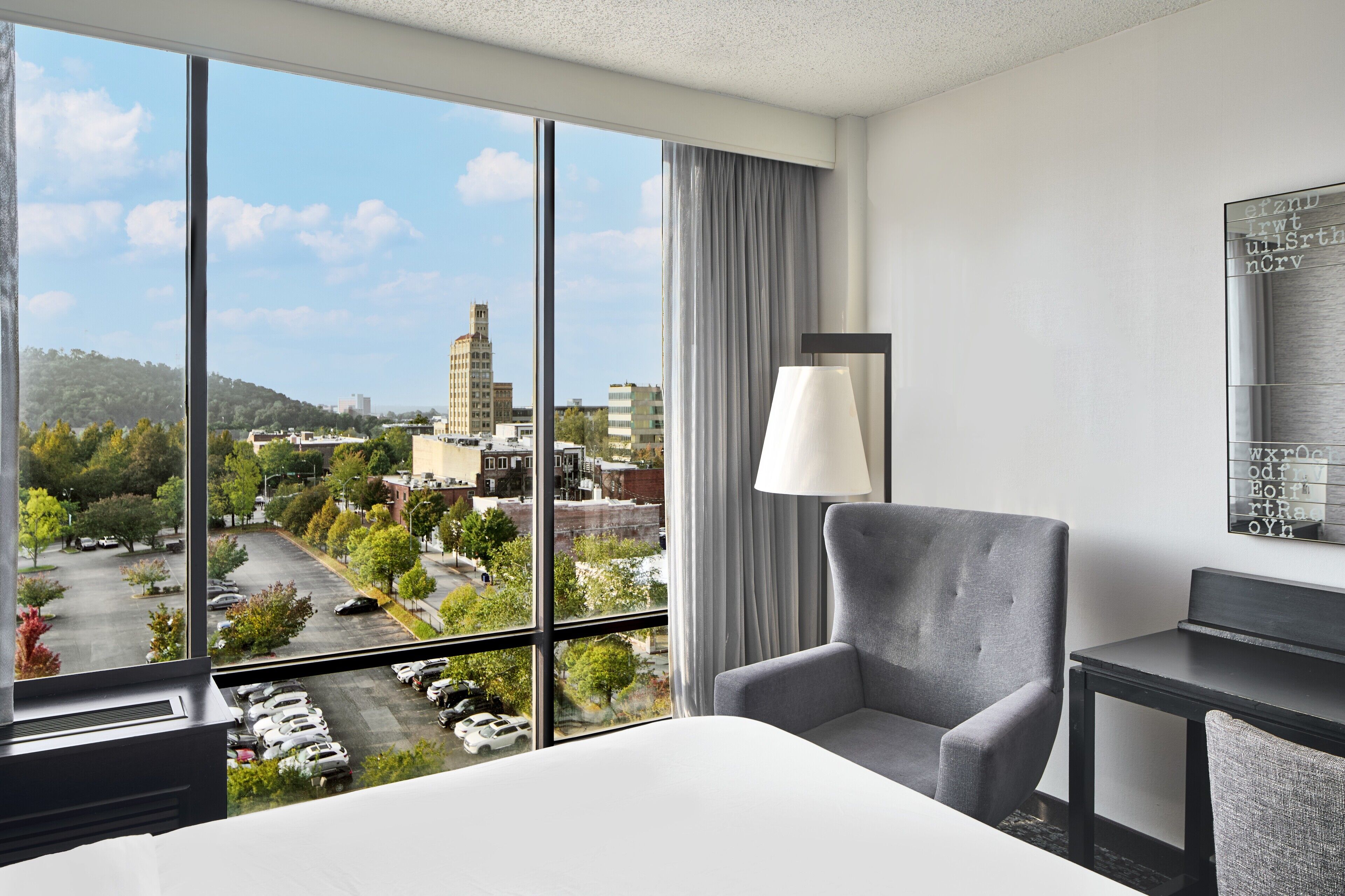 A hotel room with a large window offering a city view, a grey armchair, a bedside table, and a lamp.