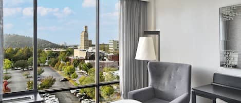 A hotel room with a large window offering a city view, a grey armchair, a bedside table, and a lamp.