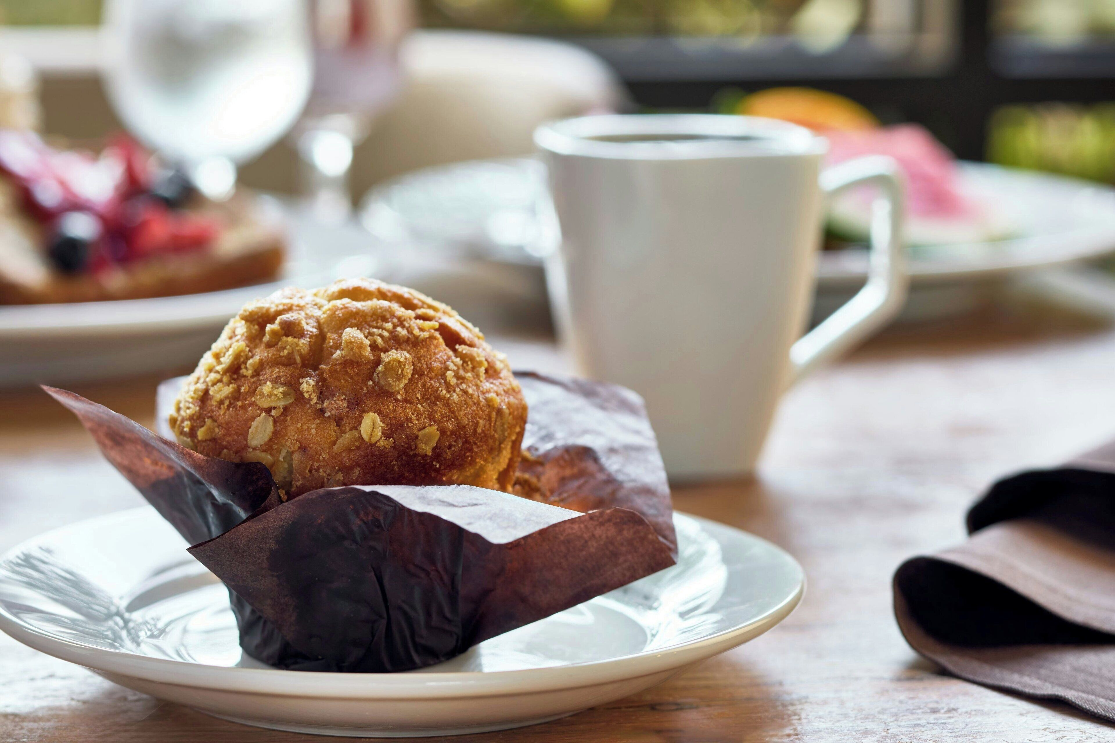 A muffin in a paper wrapper on a plate, a white cup, and a blurred background with a table.