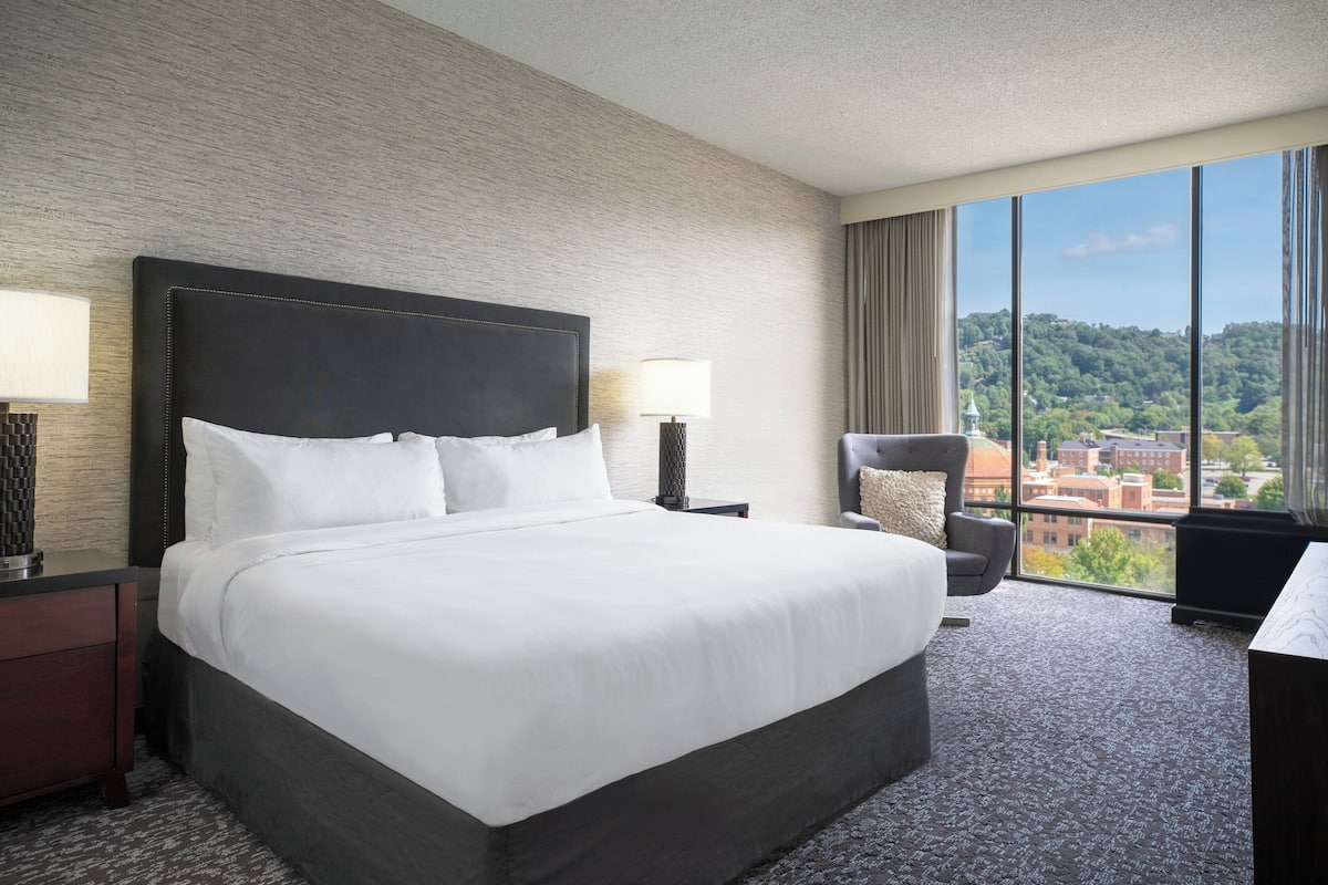 A hotel room with a large bed, a nightstand, a chair, and a view of the city through large windows.