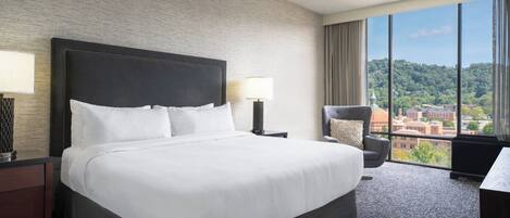 Camera d'hotel con un letto grande, un comodino, una sedia e vista sulla città attraverso ampie finestre.