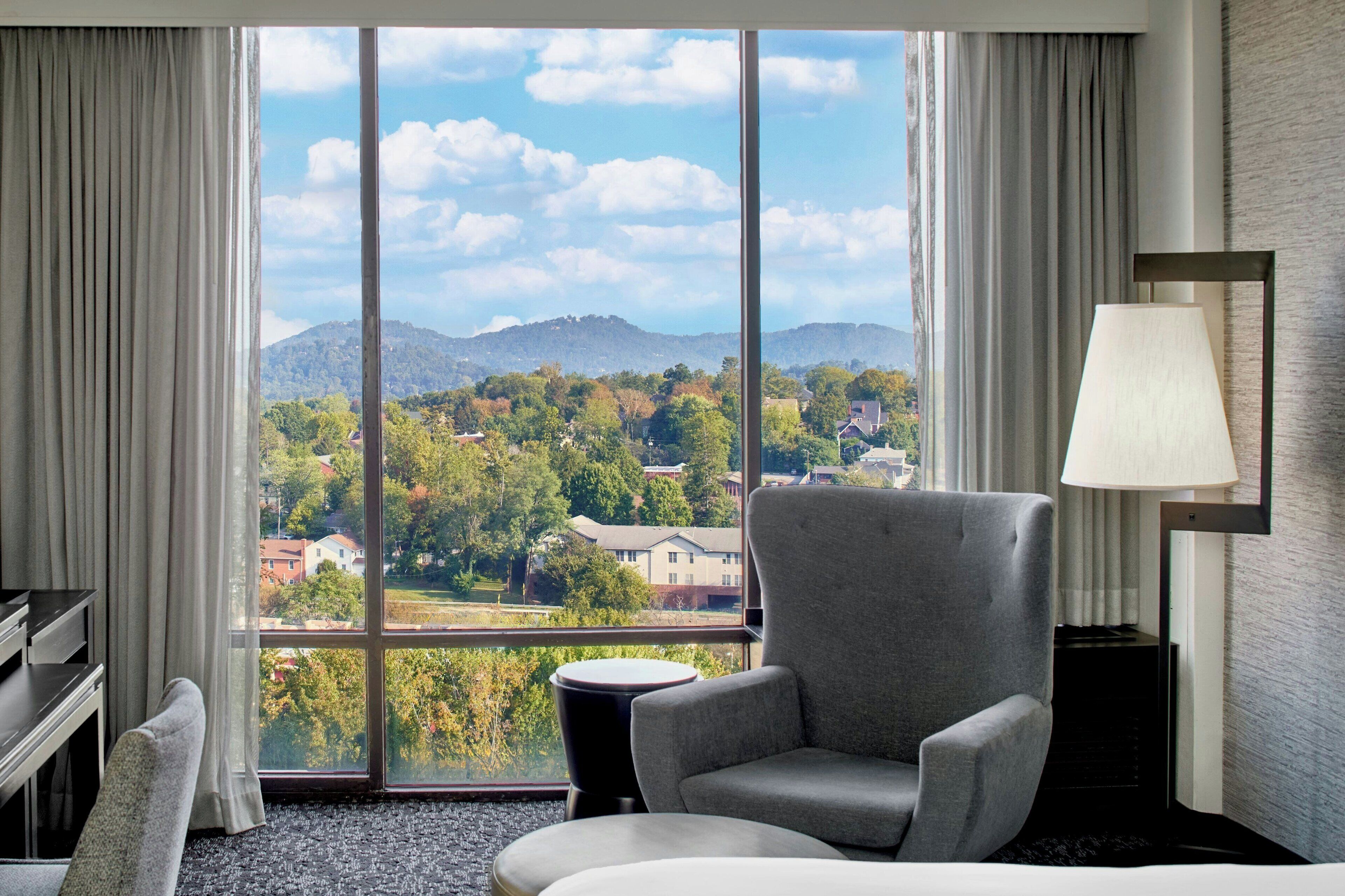 A hotel room with a large window offering a view of a mountainous landscape and residential buildings.