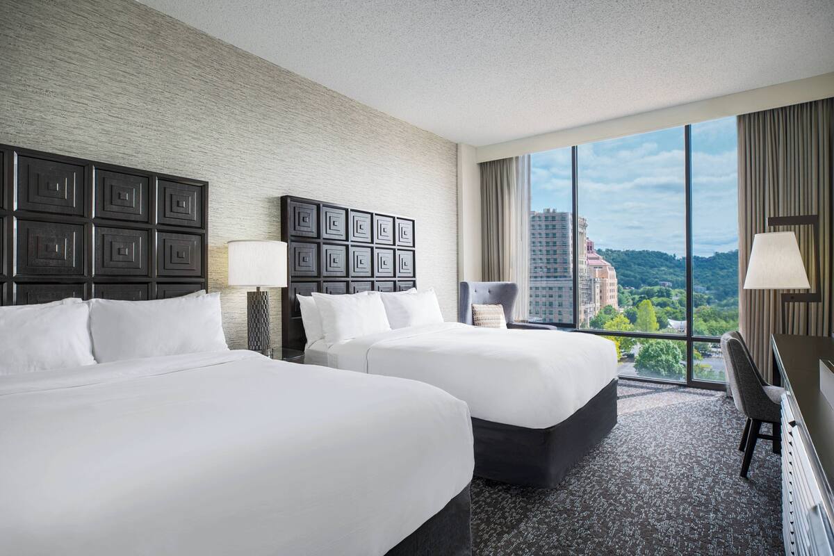 A hotel room with two beds, a large window with a city view, and a desk with a chair.