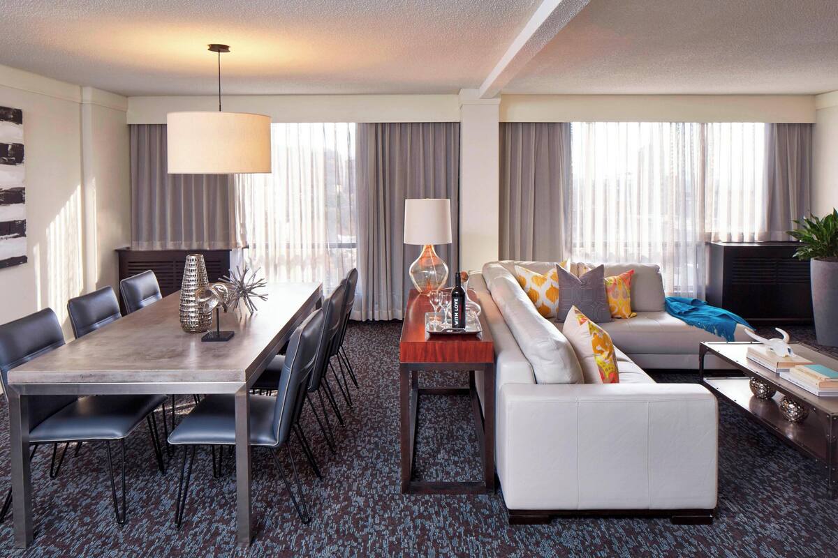 A modern hotel room with a dining area, a sofa, and a large window with curtains.