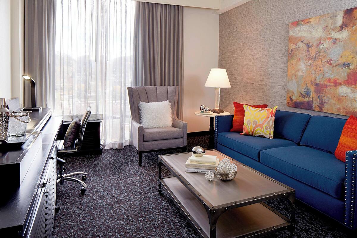 A hotel room with a grey armchair, a blue sofa, a coffee table with books, a lamp, and a large abstract painting on the wall.