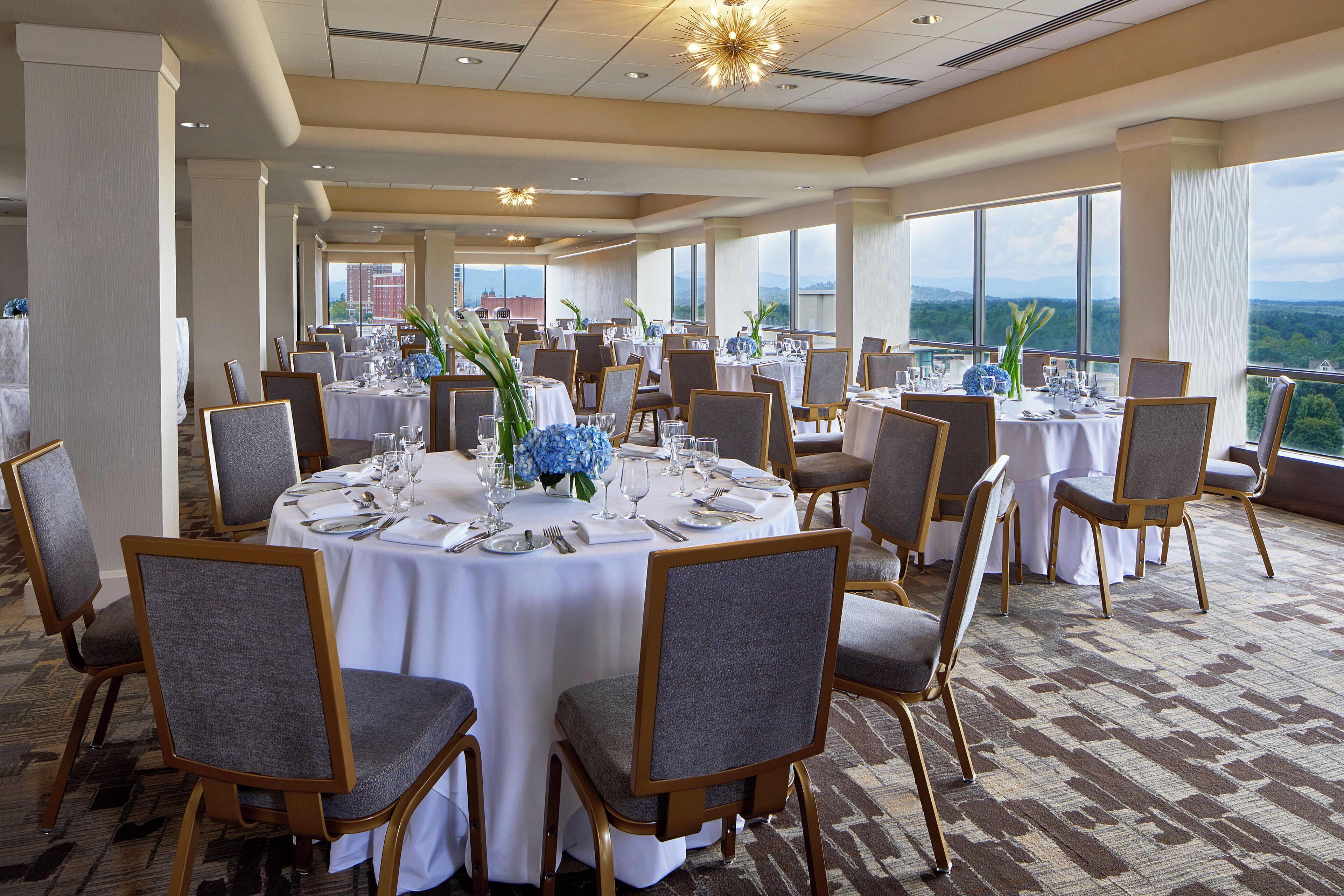 Un salón de banquetes con mesas redondas, sillas grises tapizadas y vistas a las montañas a través de amplios ventanales.