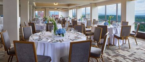 A banquet hall with round tables, grey upholstered chairs, and a view of the mountains through large windows.