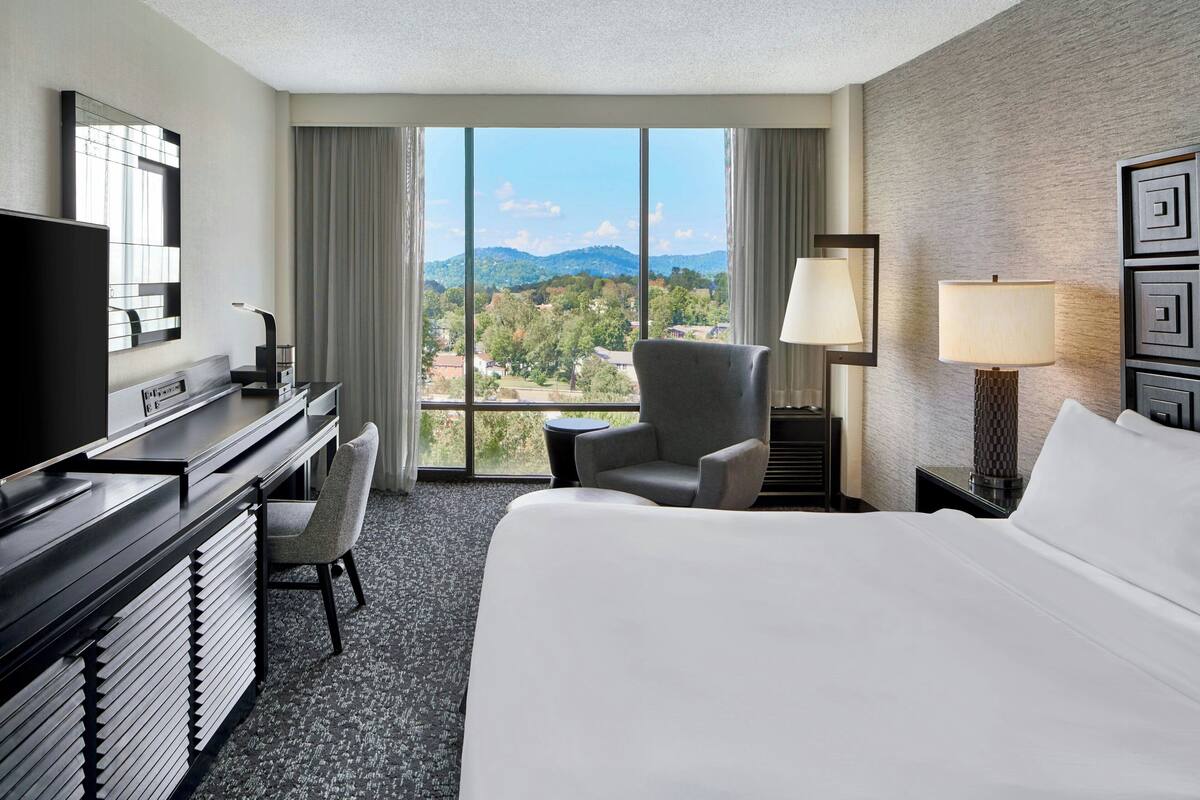 A hotel room with a large bed, a desk with a computer, a chair, a lamp, and a view of the mountains through the window.