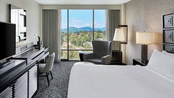 Une chambre d’hôtel avec un grand lit, un bureau avec un ordinateur, une chaise, une lampe et une vue sur les montagnes par la fenêtre.