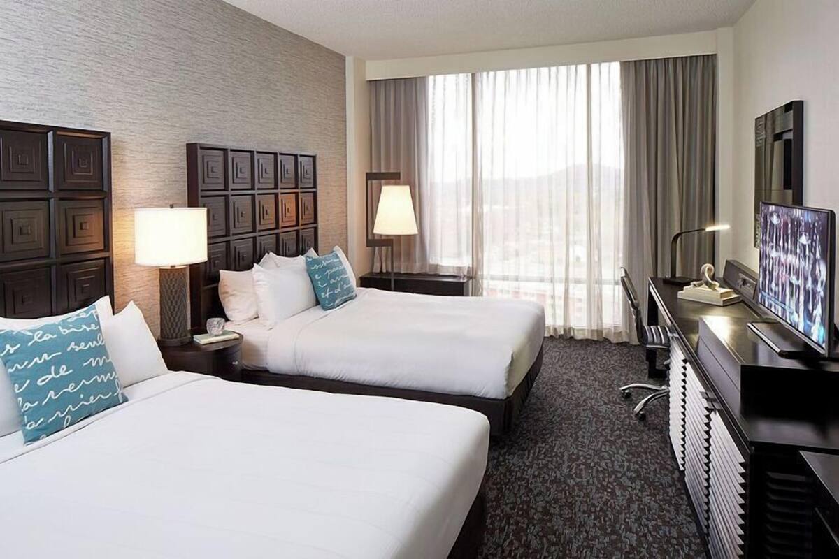 A hotel room with two beds, a desk with a TV, and a large window with curtains.