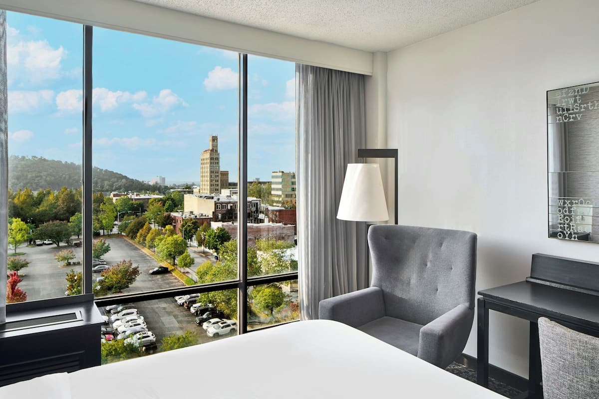 A hotel room with a large window offering a city view, a grey armchair, a bedside table, and a lamp.