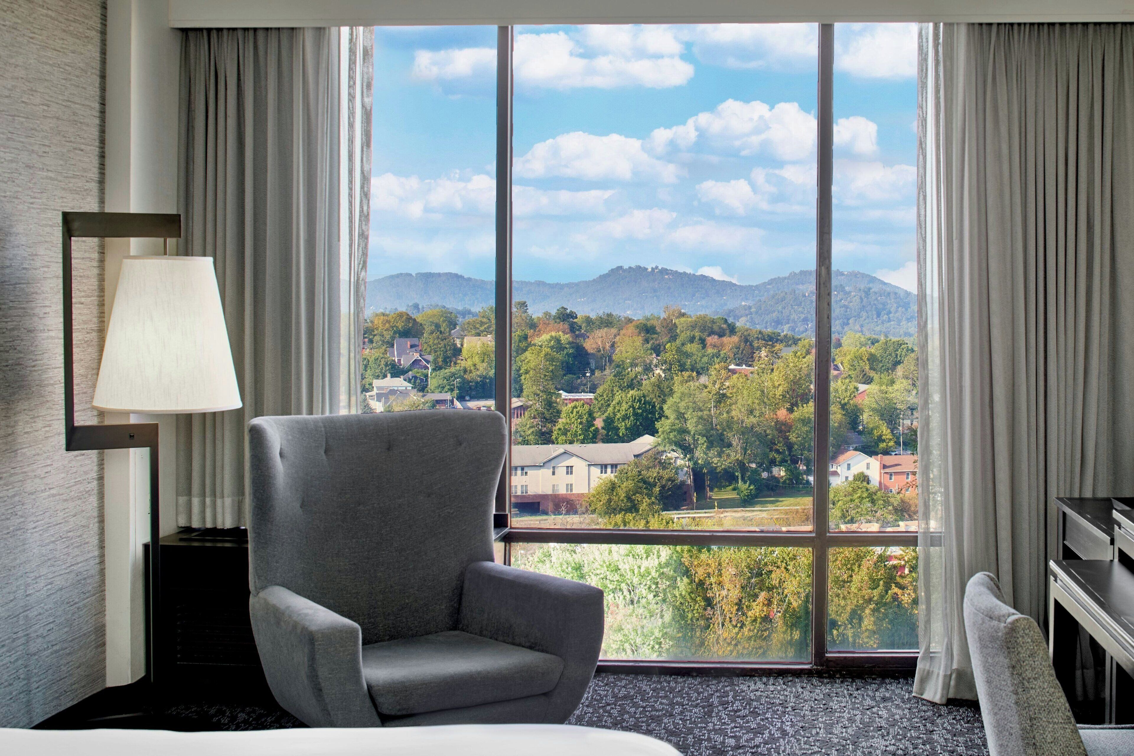 Une chambre d’hôtel avec une grande fenêtre, un fauteuil gris, un lit et une lampe.