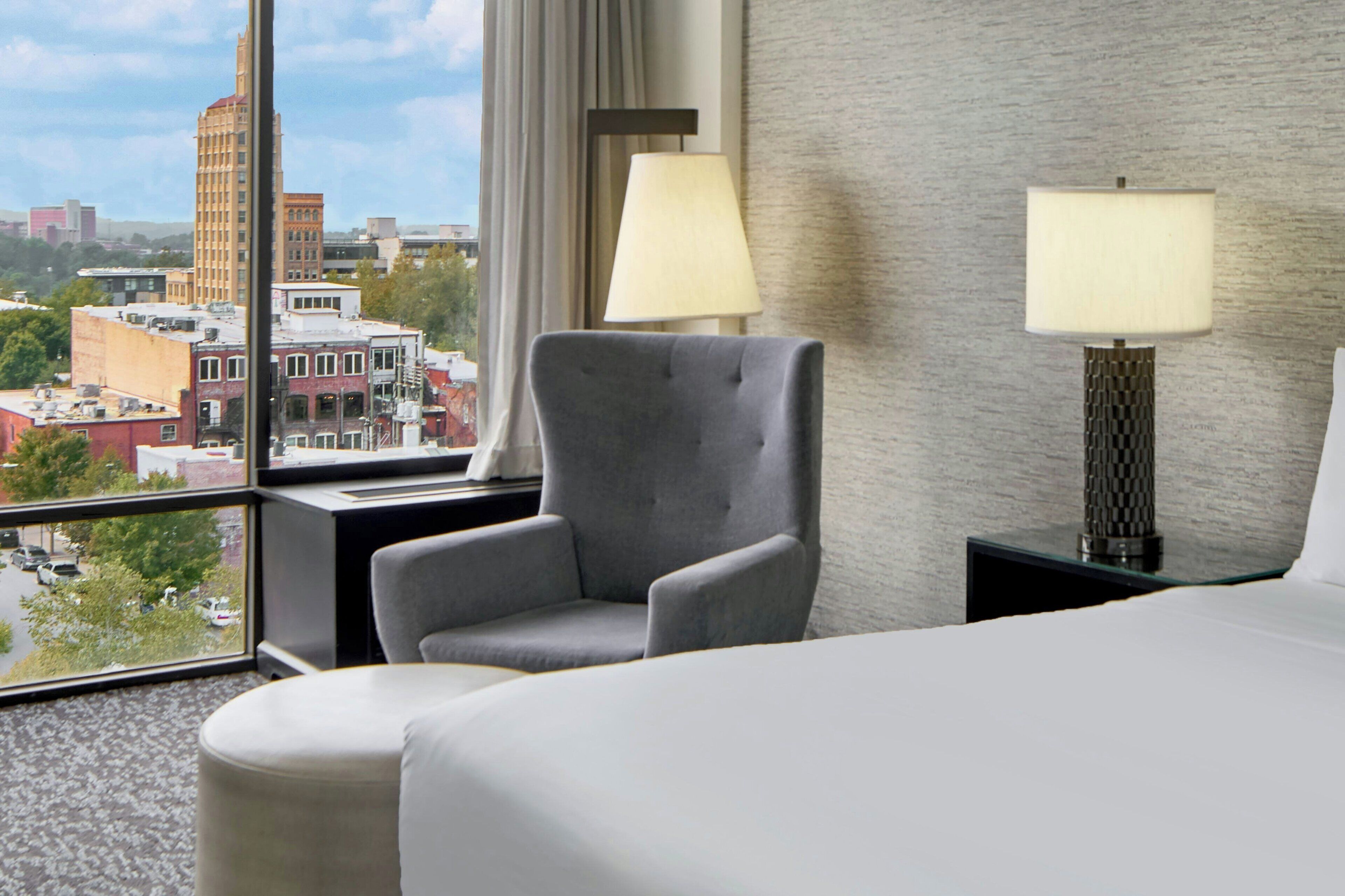 Habitación de hotel con una ventana grande que ofrece vistas a la ciudad, un sillón gris, dos lámparas de noche y una cama bien hecha.