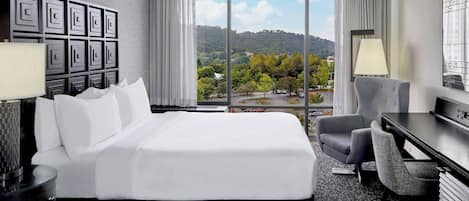 A hotel room with a large bed, a desk, a chair, and a view of a green landscape through large windows.