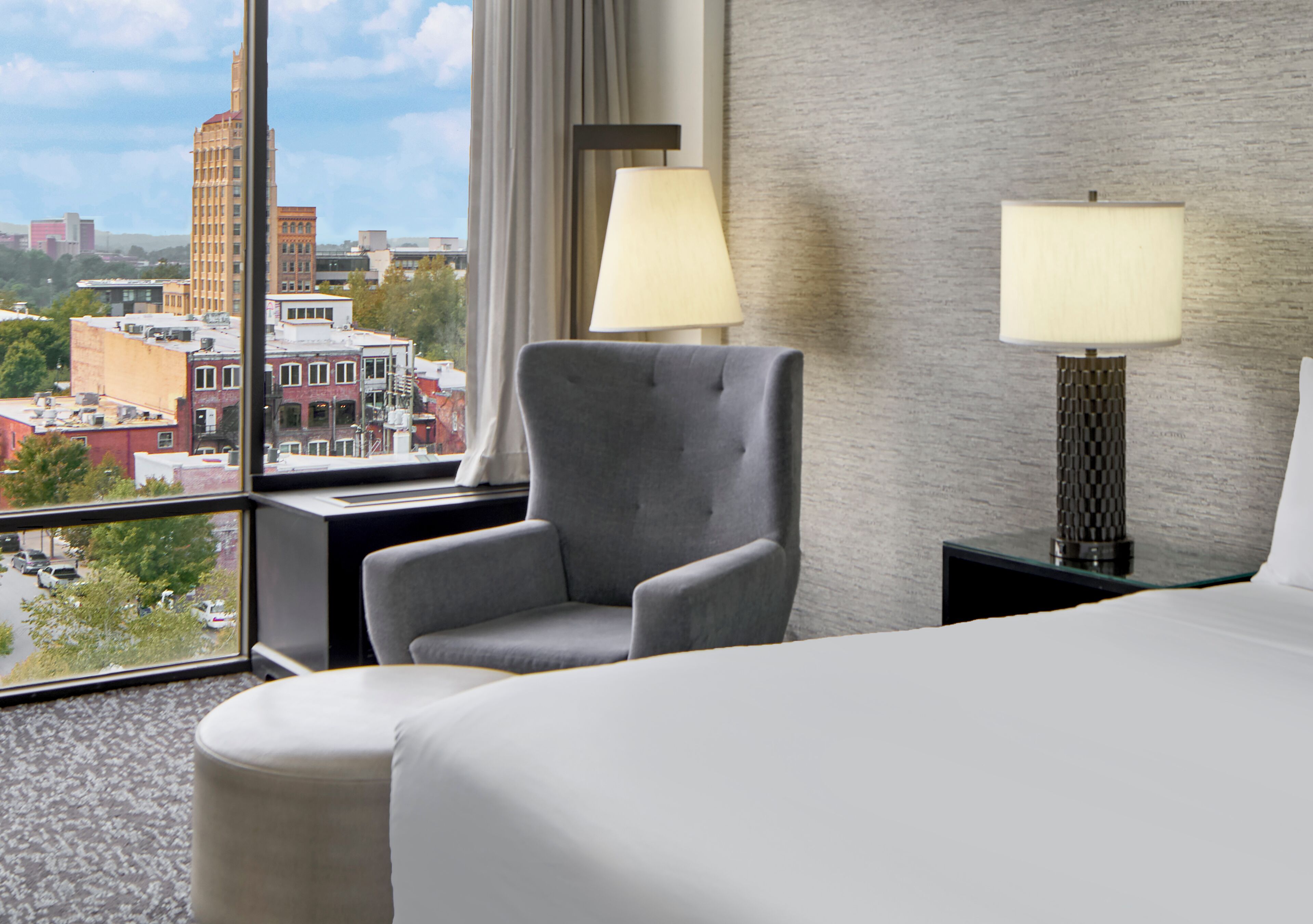 Ett hotellrum med ett stort fönster som vetter mot staden, en grå fåtölj, en säng med vita sängkläder och två sänglampor.