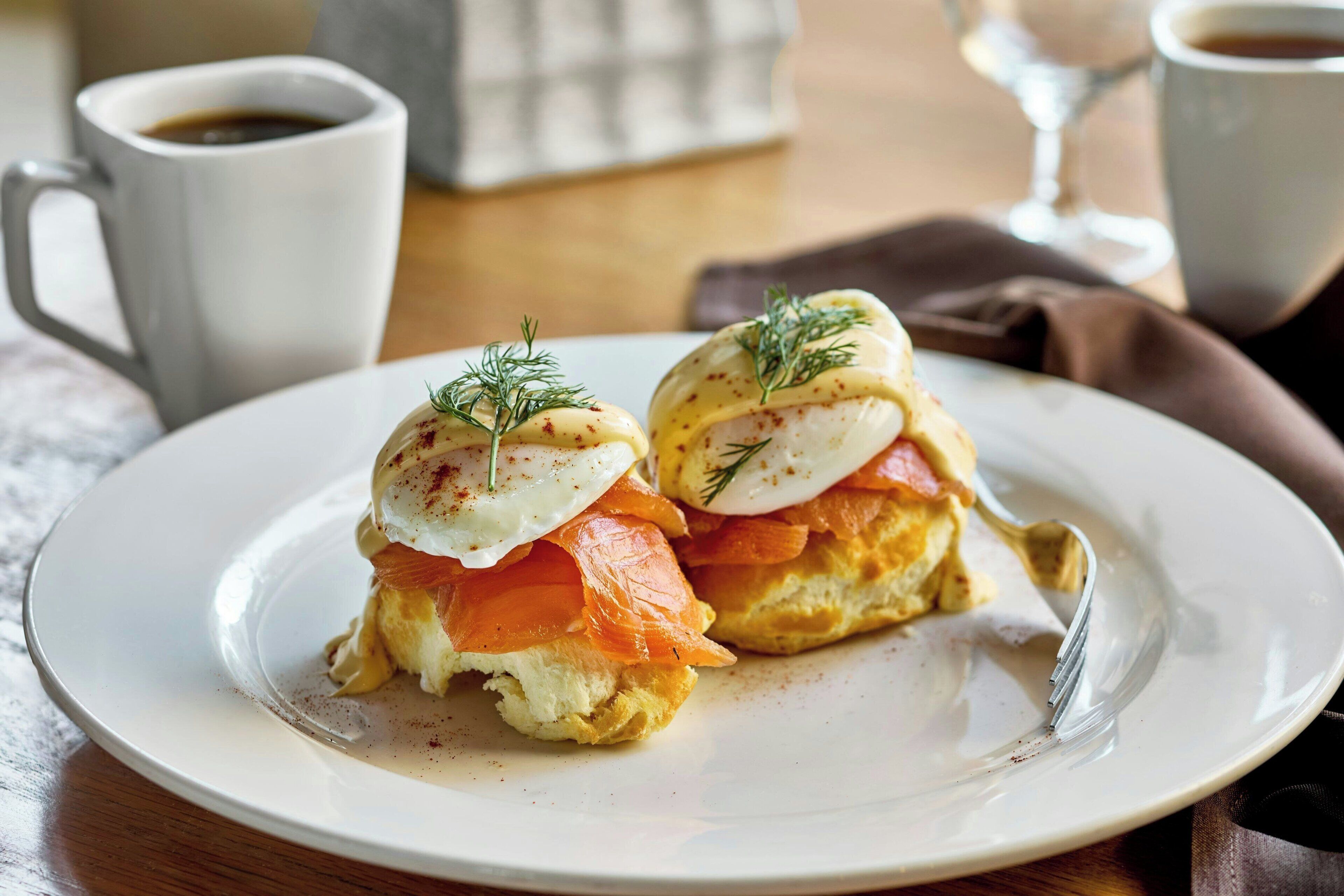 A plate with two eggs benedict sandwiches, a fork, and two cups of coffee on a table.