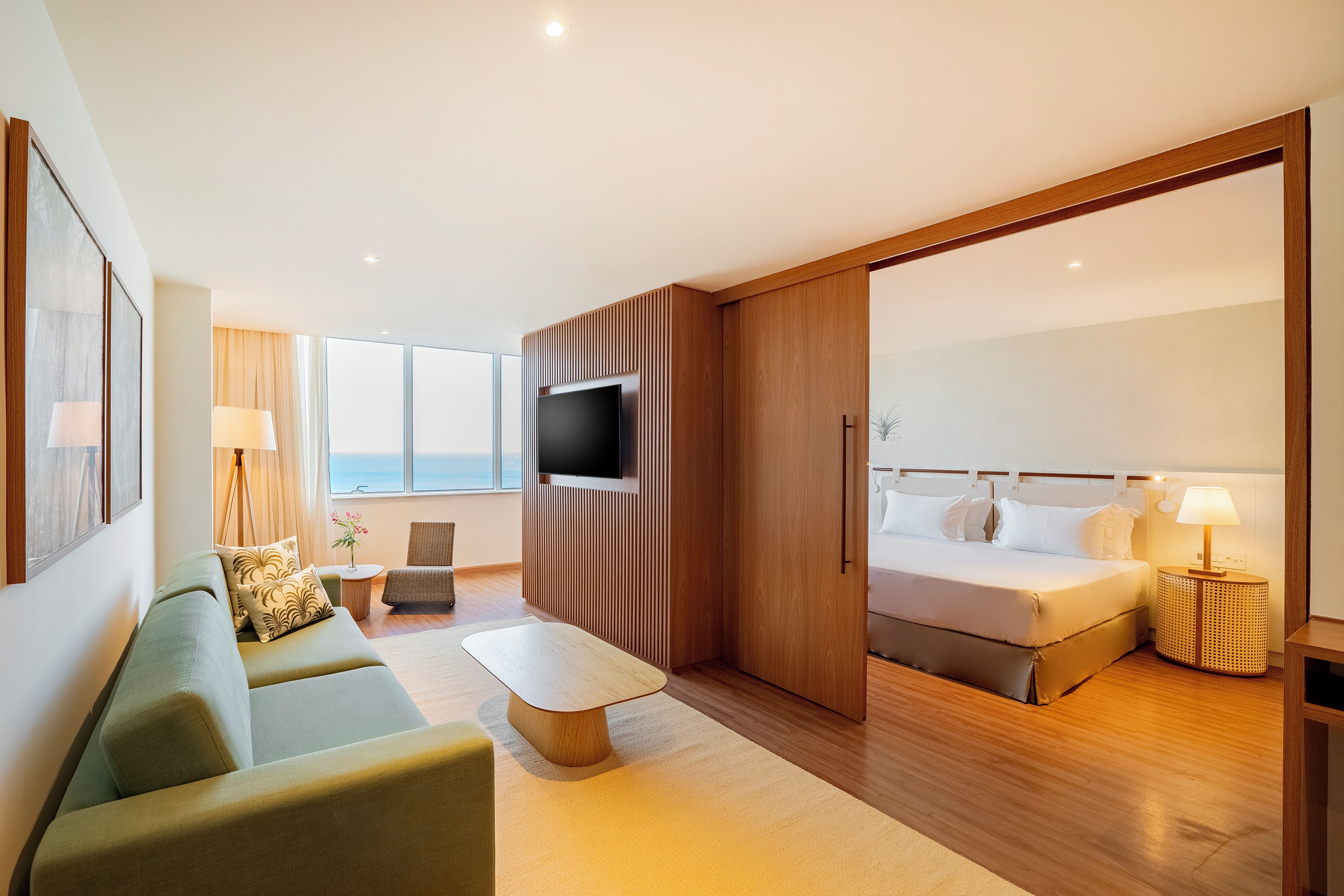 A modern hotel room with a sofa, a TV, a bed, and a view of the sea.