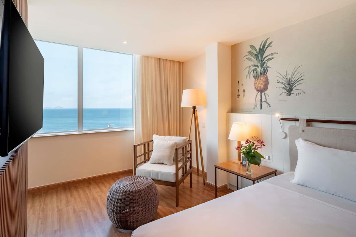 A hotel room with a bed, a chair, a small table with a lamp, a round ottoman, a television, and a large window with a view of the sea.